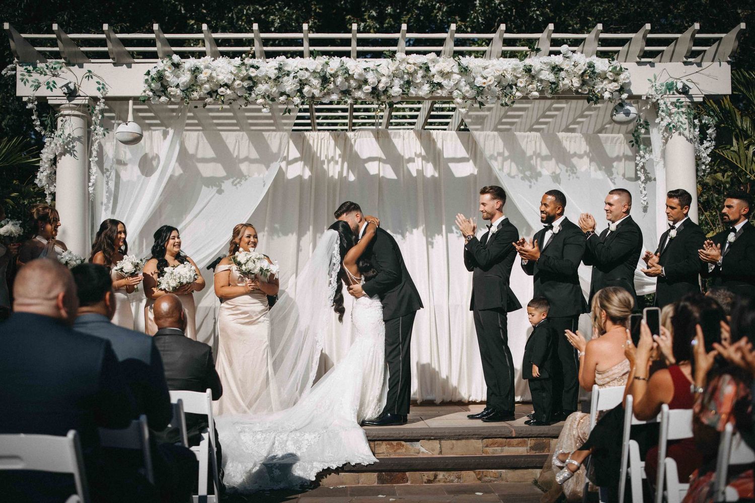 Bride and groom share their first kiss under a floral arch surrounded by wedding party and guests.