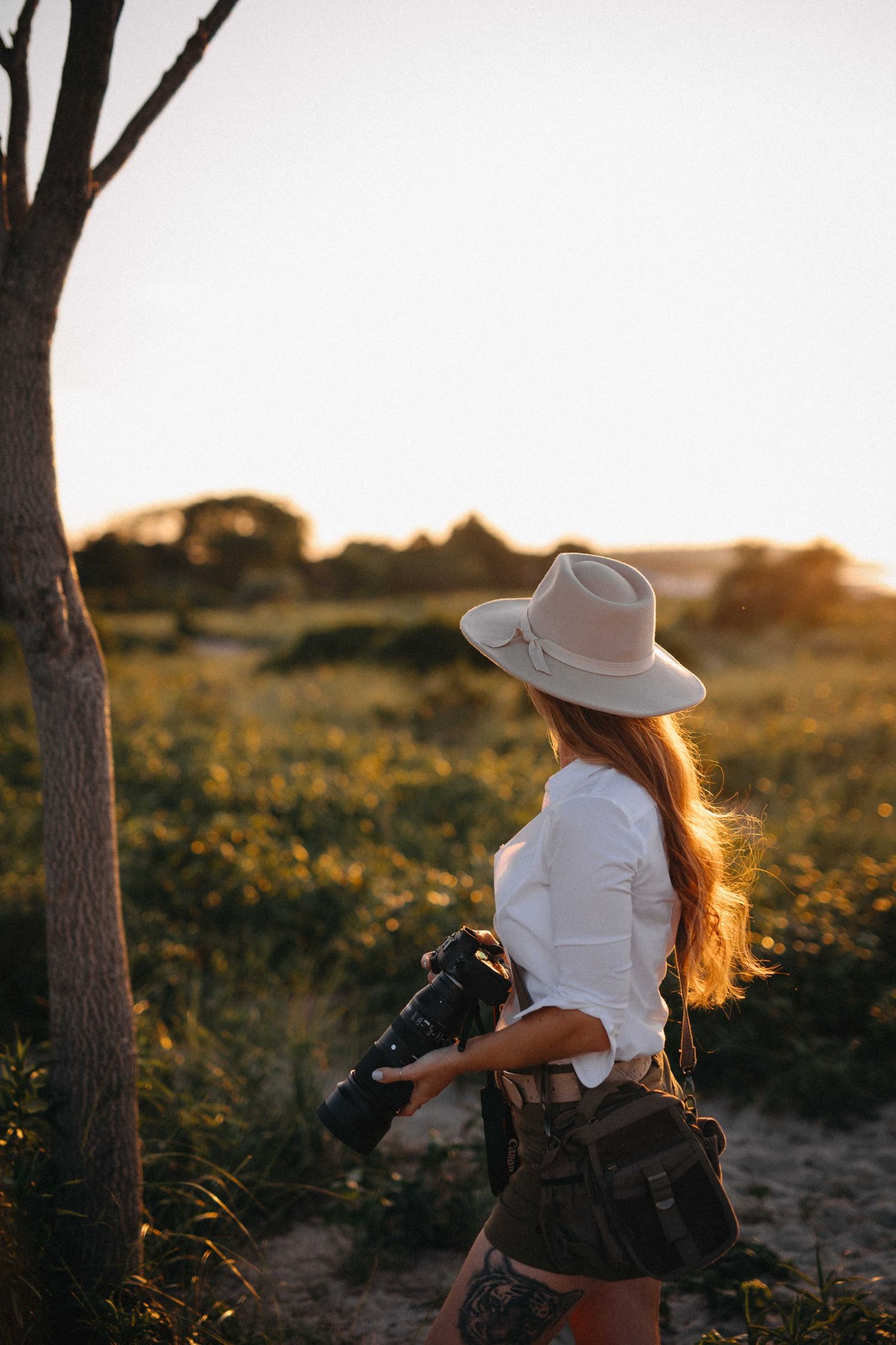 Woman in a wide-brim hat and white shirt holding a camera while walking through golden sunlit fields.