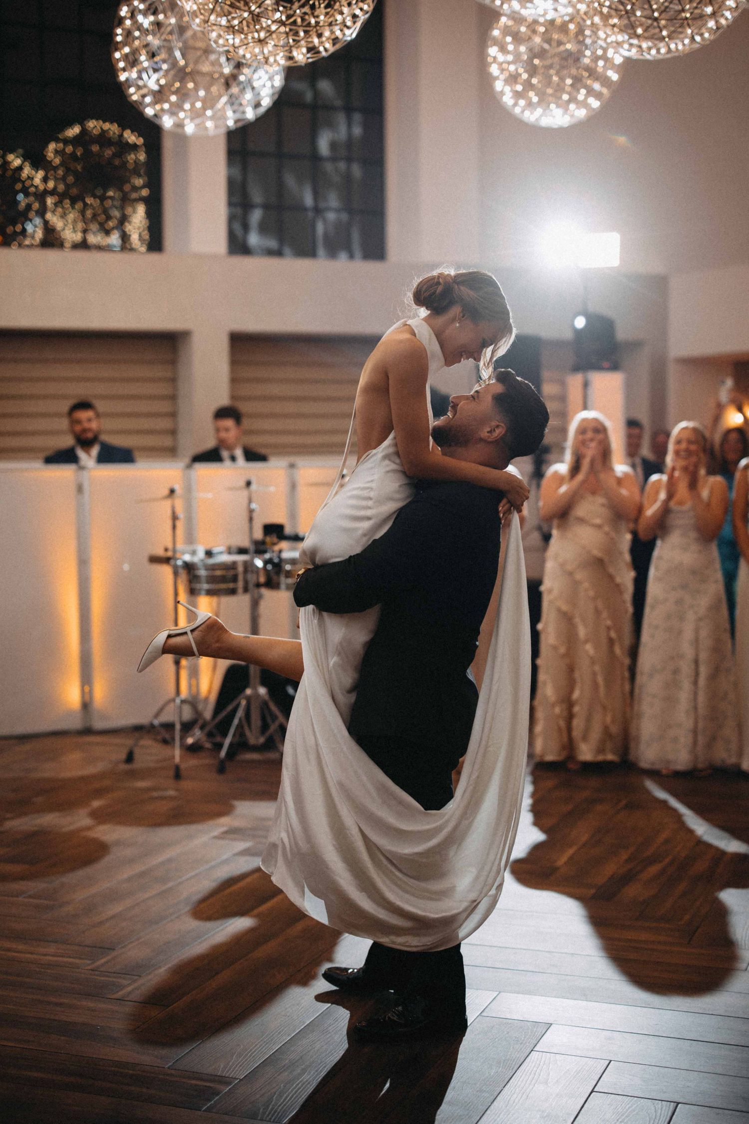 Groom lifts bride during emotional first dance under sparkling chandeliers at reception.