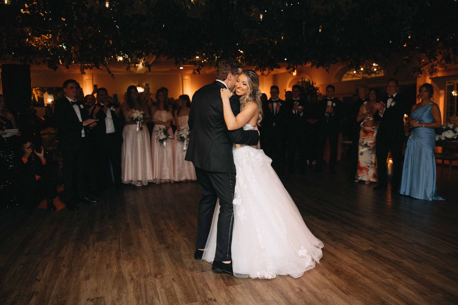 Bride and groom sharing romantic first dance surrounded by guests in warmly lit venue.