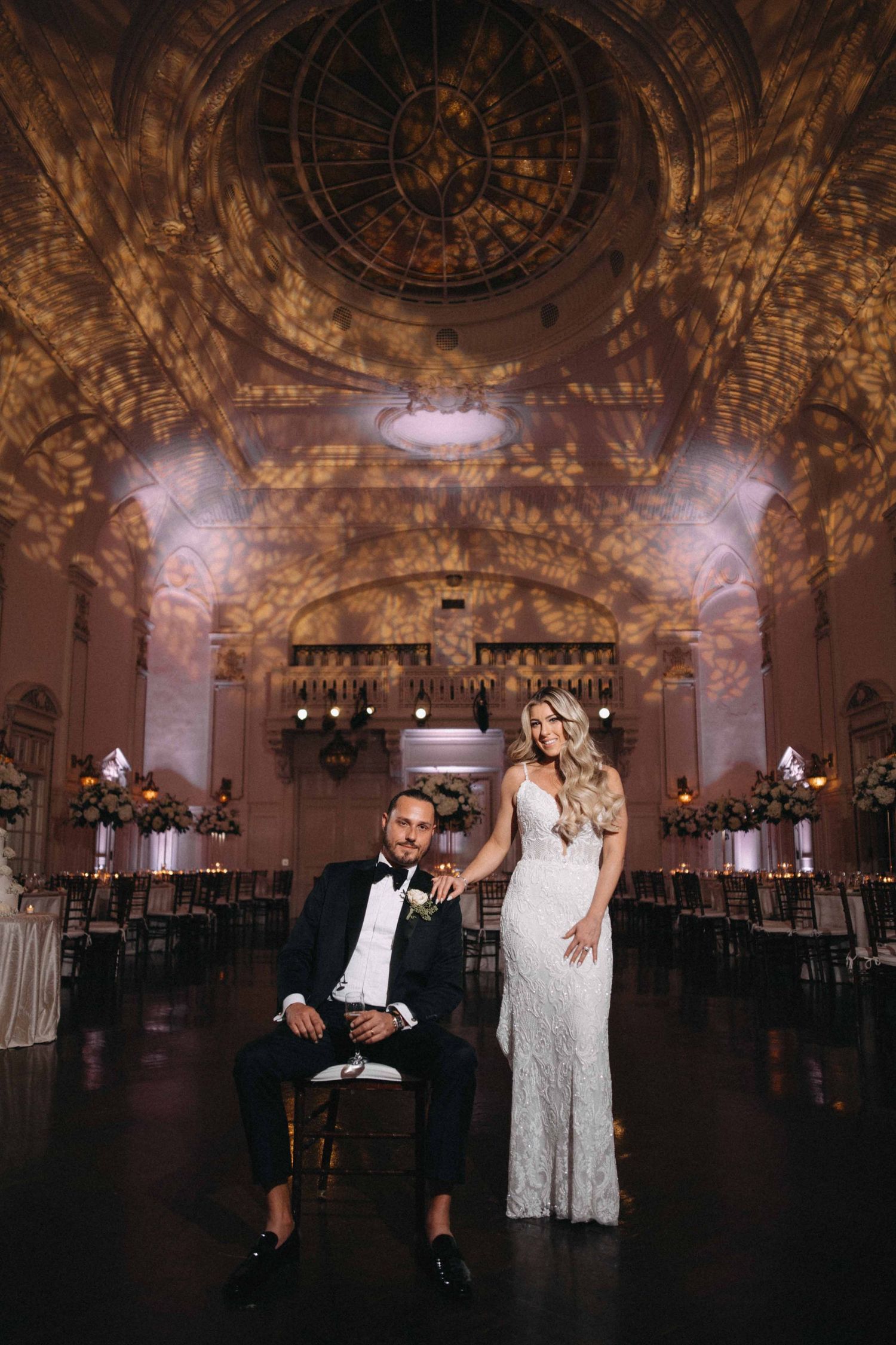 Bride in lace gown and groom in tuxedo posing in ornate ballroom with stunning golden ceiling projections.
