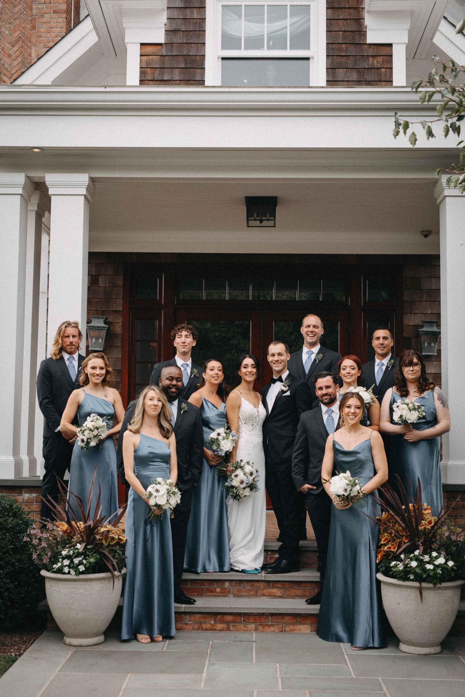 Wedding party in dusty blue gowns and black tuxedos posing on steps of a craftsman-style home.