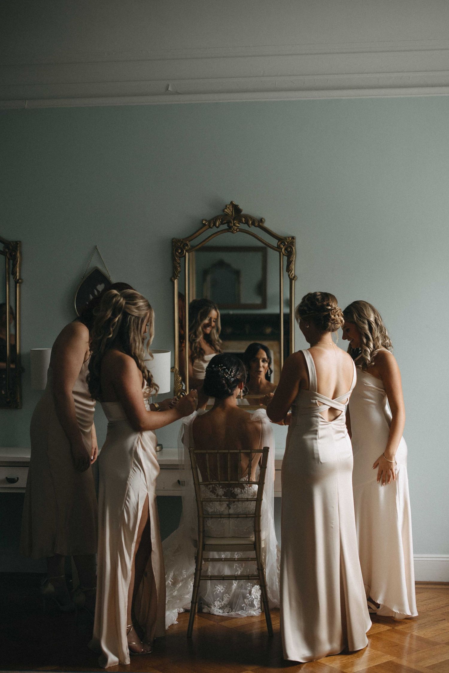 Bride seated at a vanity mirror surrounded by bridesmaids in champagne gowns in an elegant room.