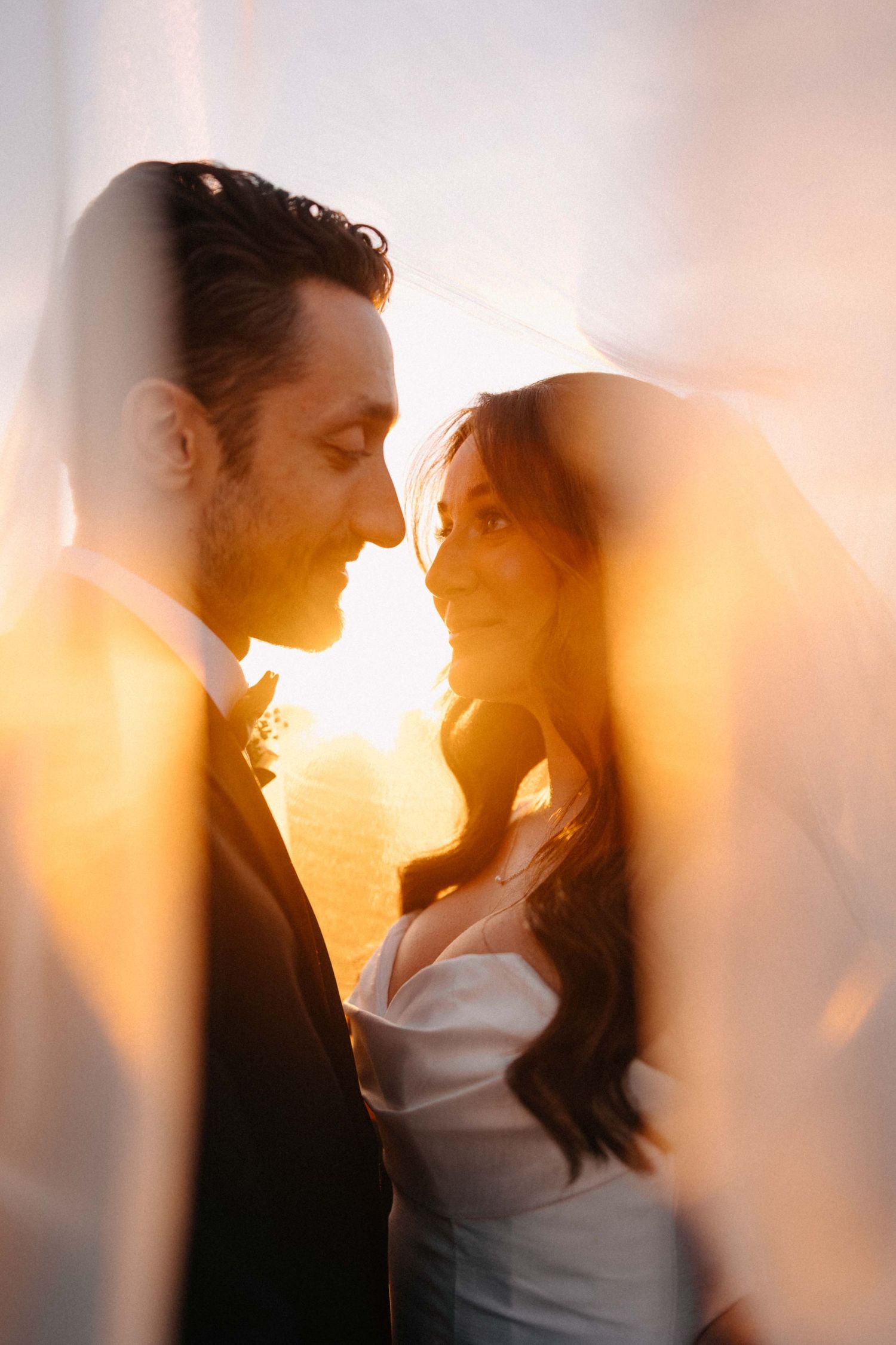 Romantic bride and groom portrait through sheer veil with golden sunset backlight.