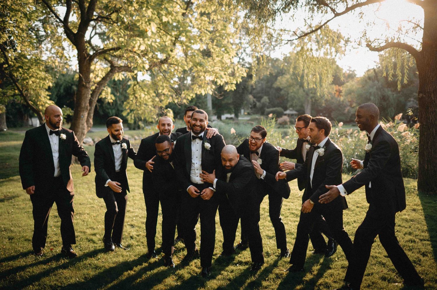 Groom and groomsmen in black tuxedos laughing and celebrating together outdoors in lush garden.