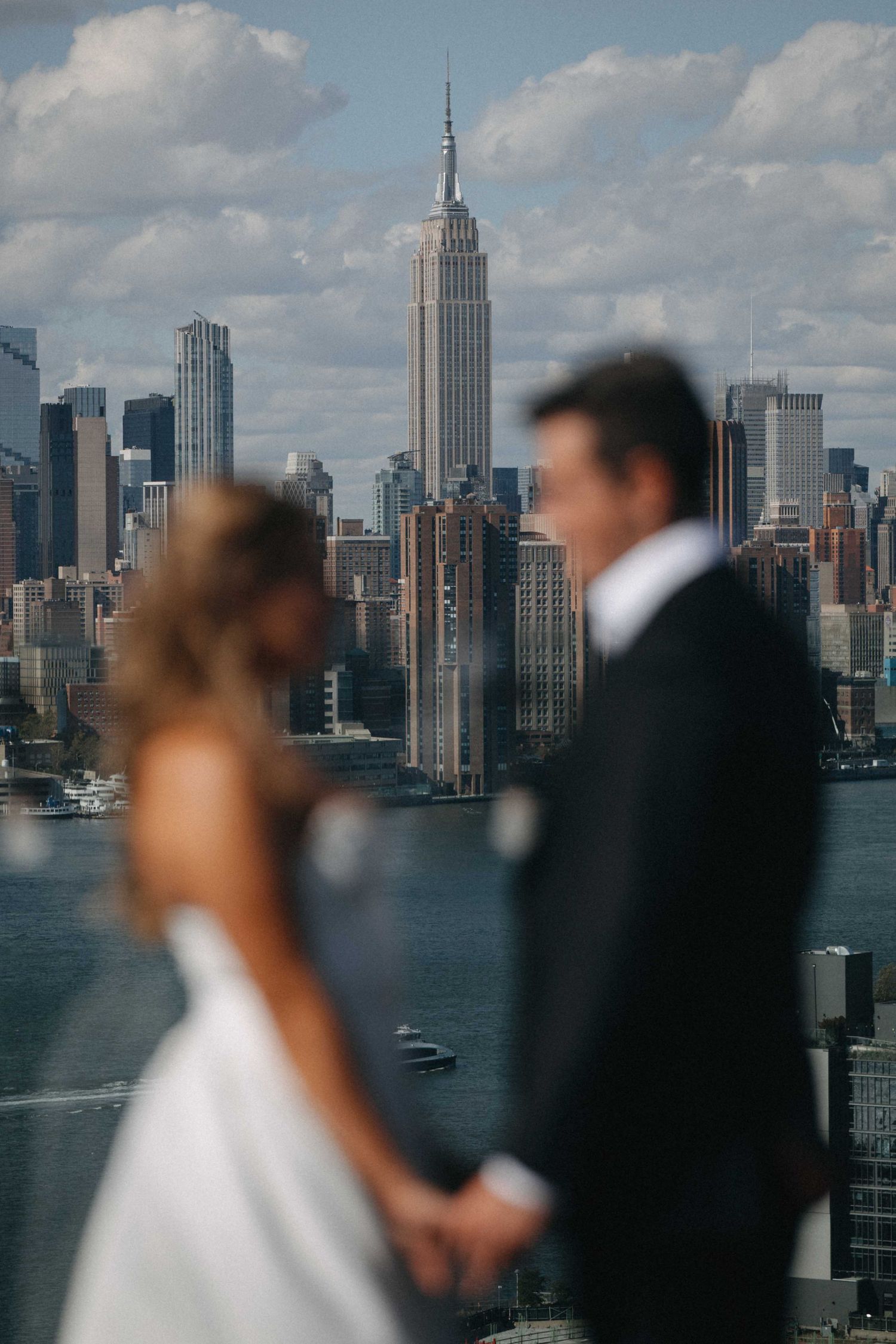 A bride and groom stand with the Empire State Building and NYC skyline visible behind them.