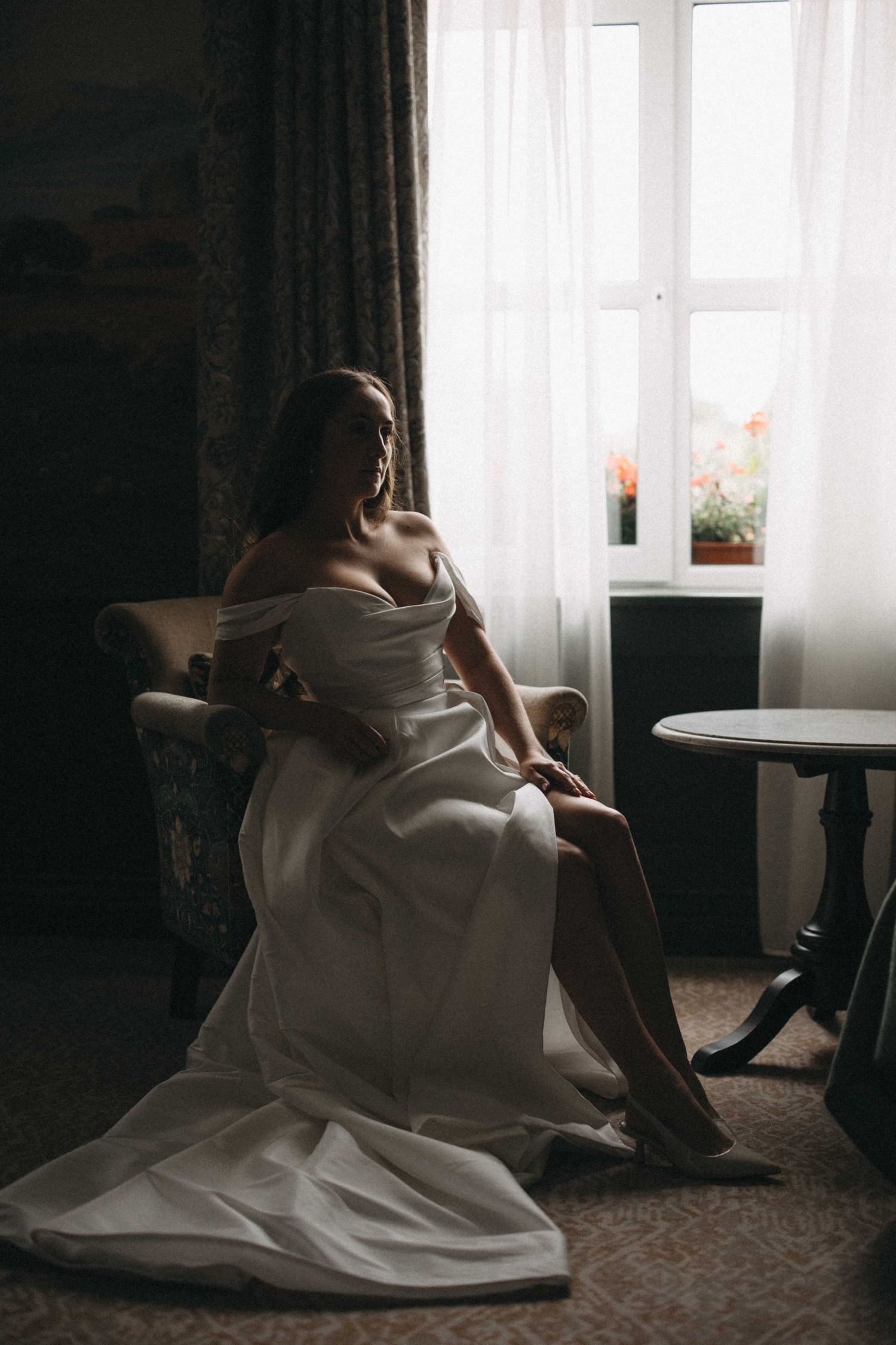 Bride in an off-shoulder white wedding gown sits elegantly in an armchair by a sunlit window in a vintage room.