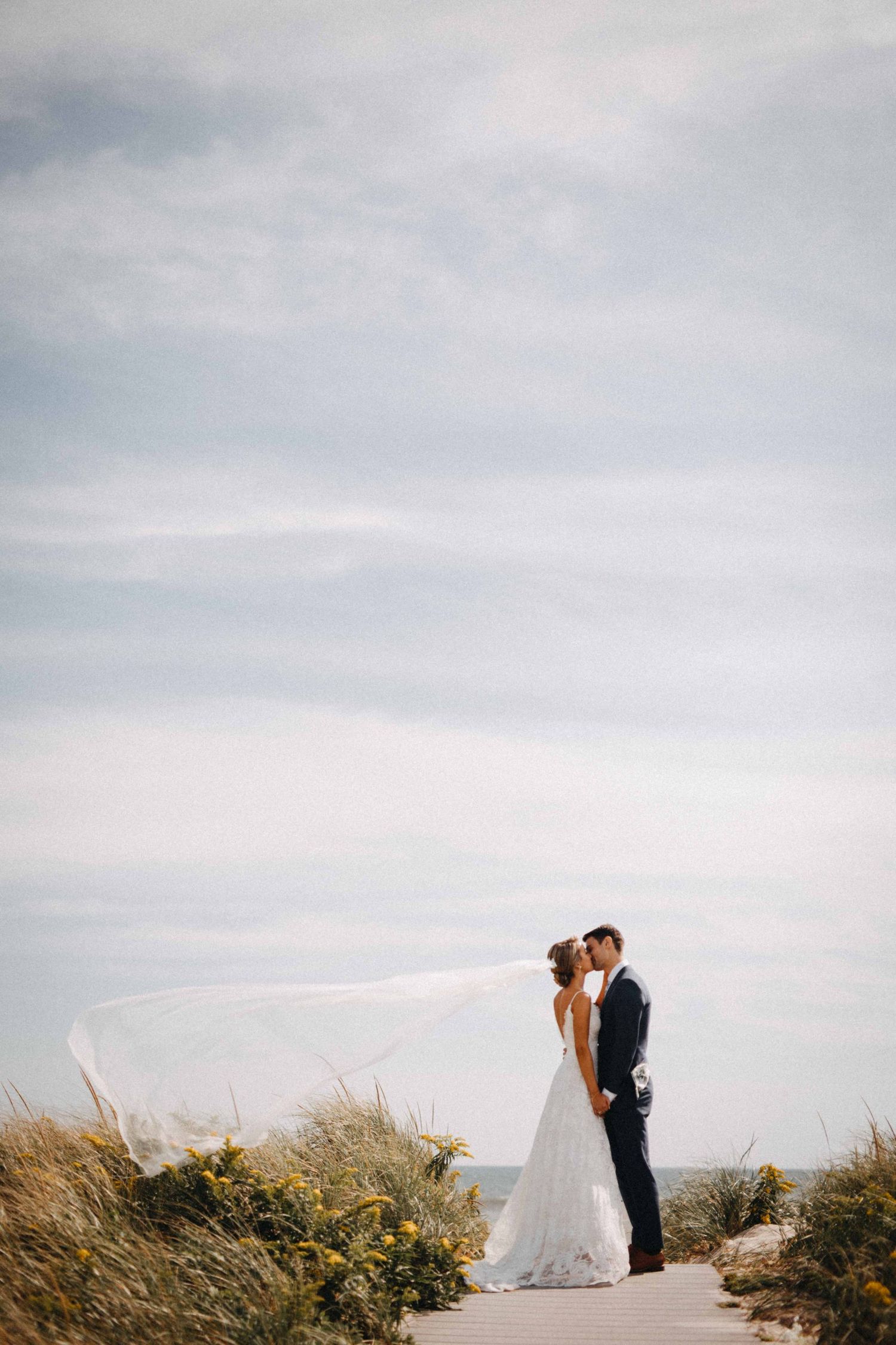 Bride and groom share romantic kiss on beach boardwalk as veil dramatically billows in wind.