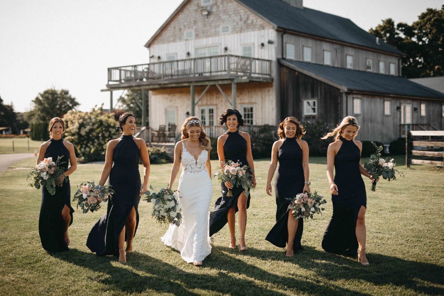 Bridal party walking in black halter gowns with bride in fitted lace dress at golden hour farm venue.