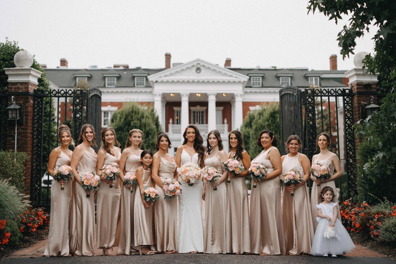 Bride with bridesmaids in champagne gowns holding pink bouquets before elegant colonial mansion gates.