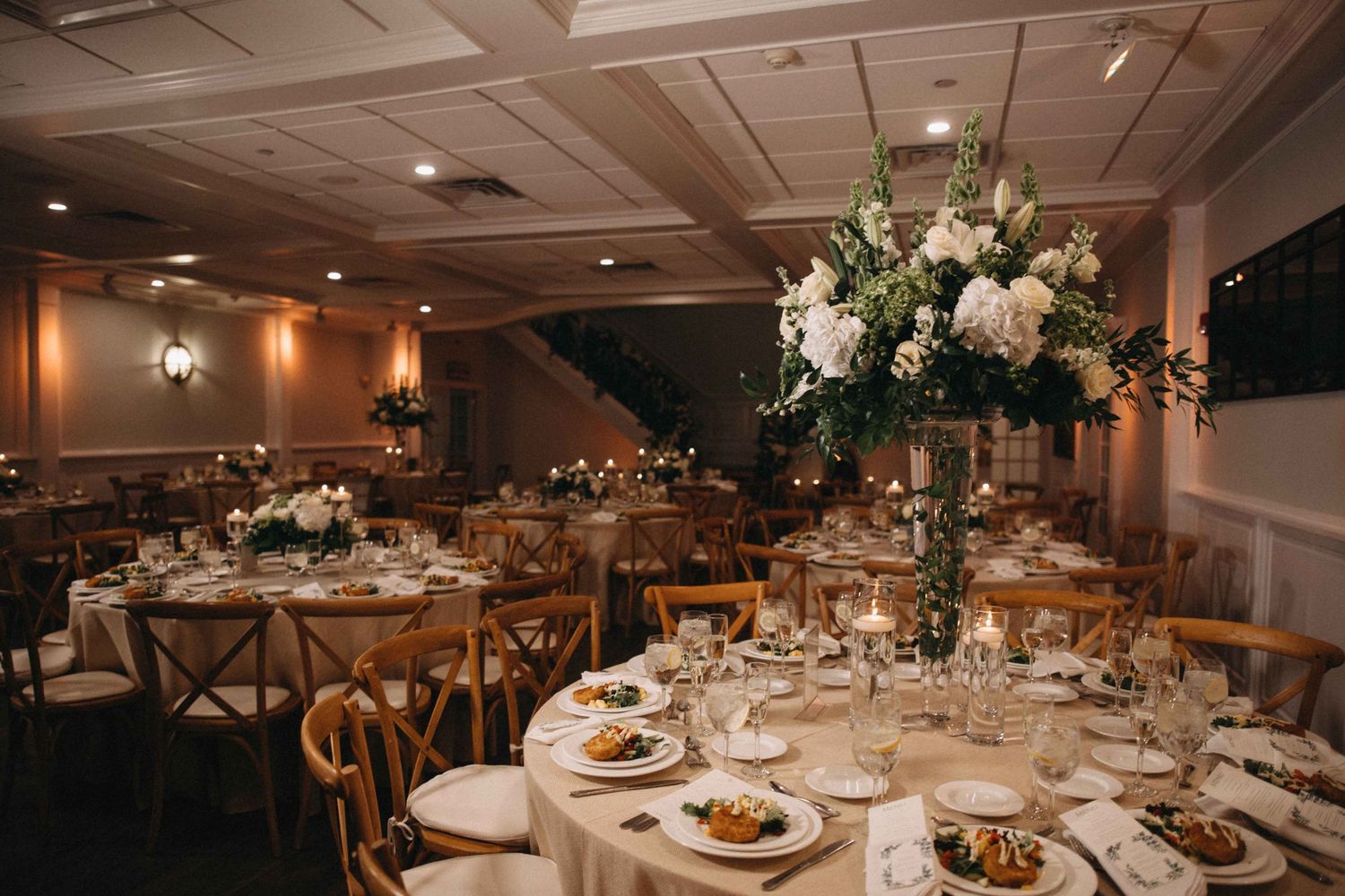 Elegant wedding banquet hall with round tables, wooden cross-back chairs, and tall white floral centerpieces with greenery.