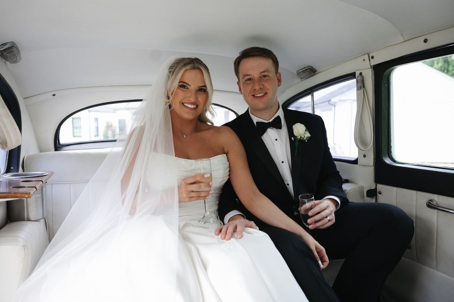 Bride and groom smiling together in a classic white wedding car, holding drinks on their special day.