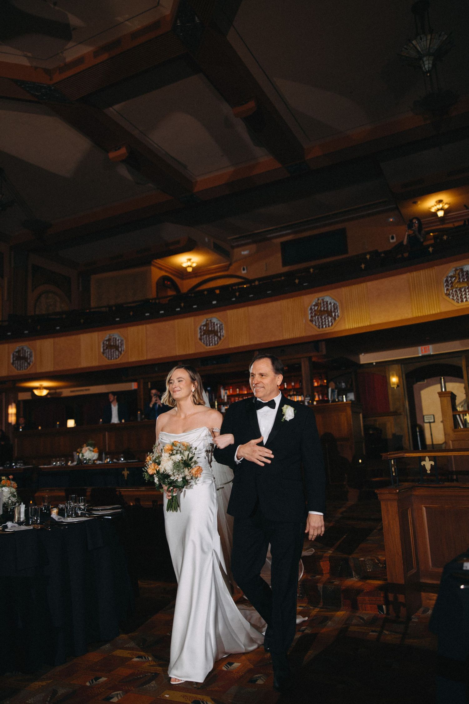 Bride in off-shoulder gown walks with father through ornate wood-paneled wedding reception hall.