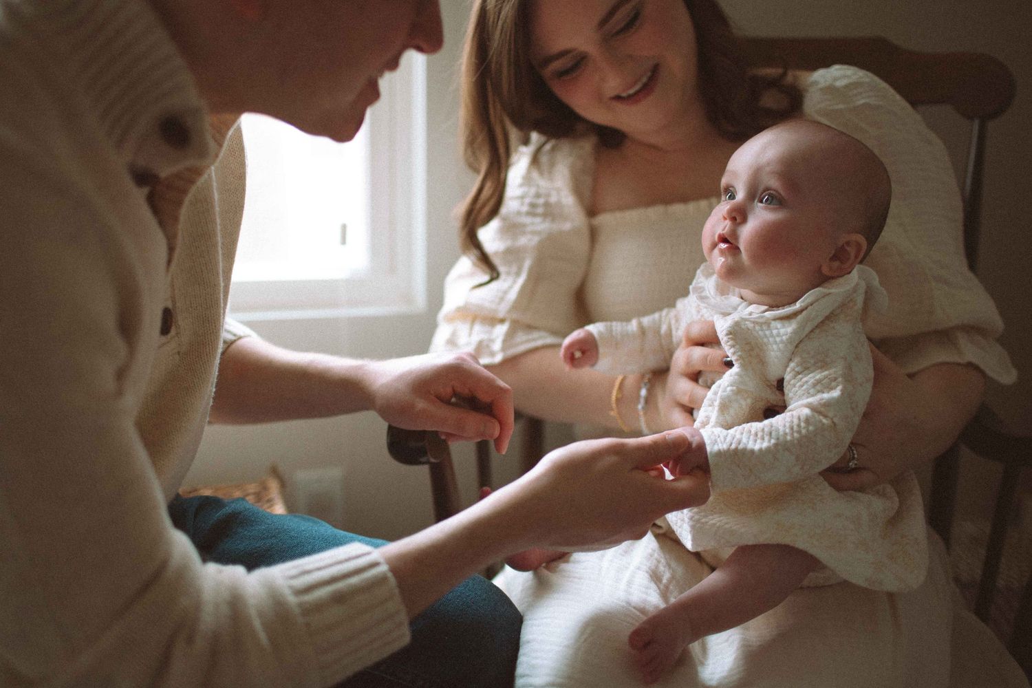 A young mother holds her newborn baby while another person gently touches the infant's hand in a warmly lit room.
