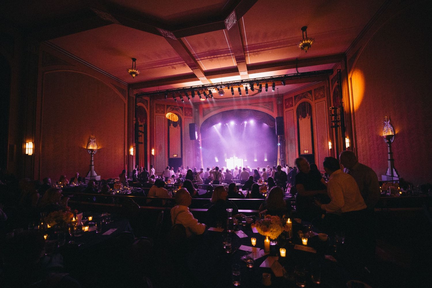 Guests enjoy a wedding reception in a stunning ornate theater venue with purple-lit stage.