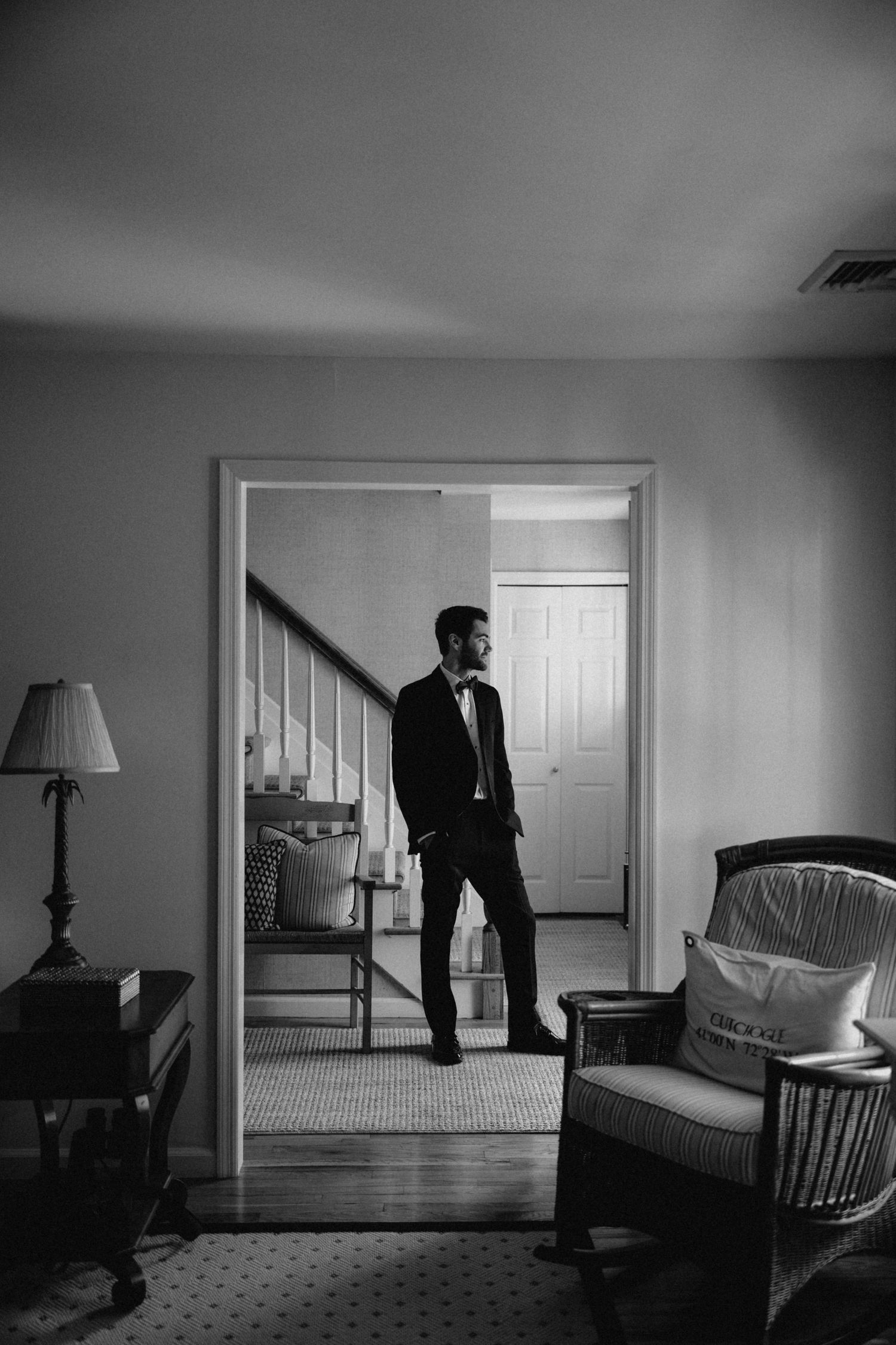 Groom in tuxedo standing in doorway of elegant home, captured in moody black and white wedding portrait.