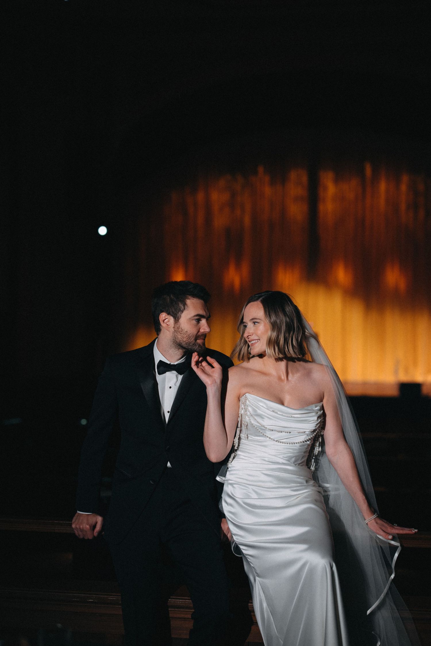 Bride and groom share romantic moment in dimly lit venue with warm golden backdrop.