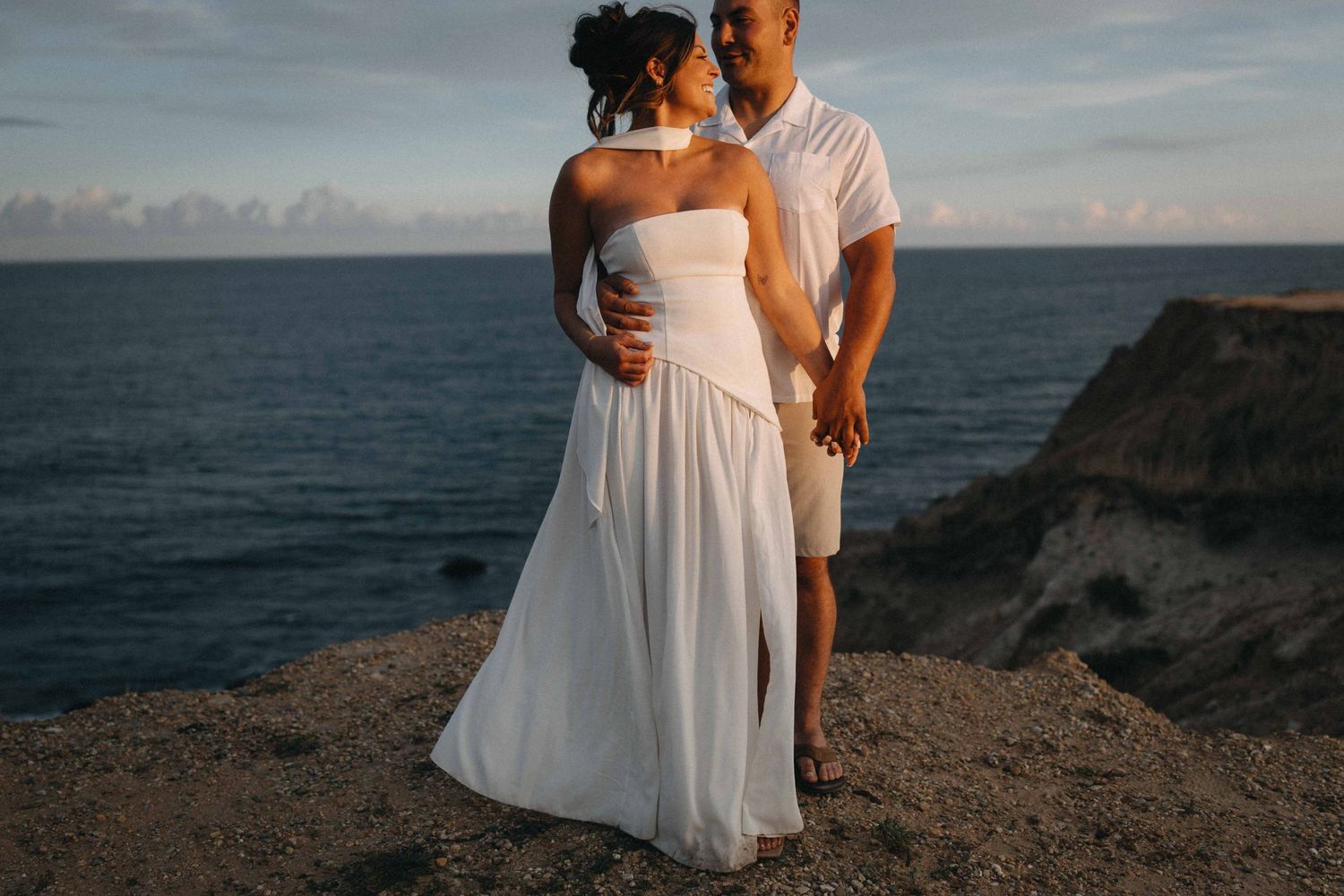 Couple in white attire standing on a rocky cliff overlooking the ocean at sunset.
