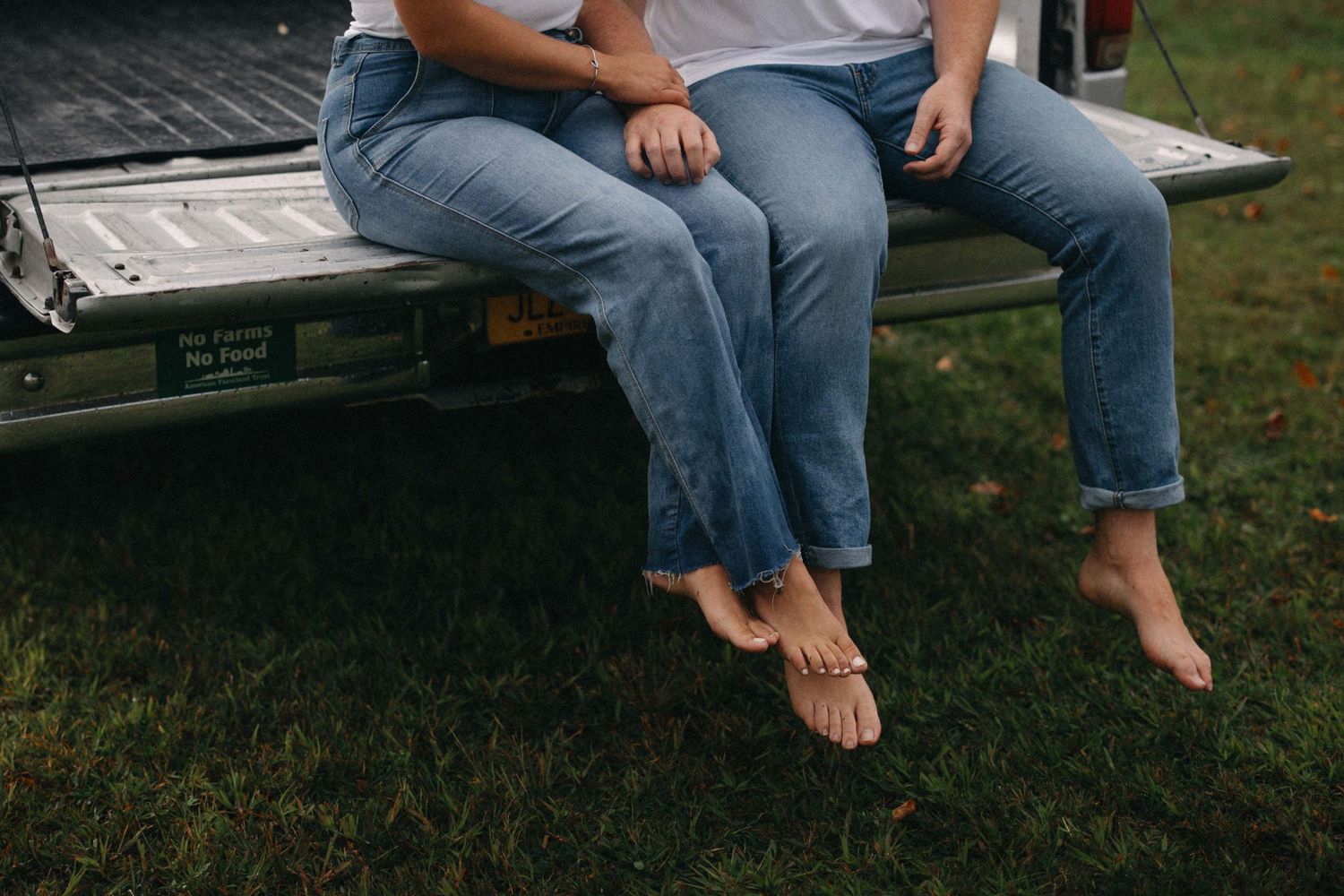 Barefoot couple sitting closely together on a truck tailgate, holding hands in a grassy field.