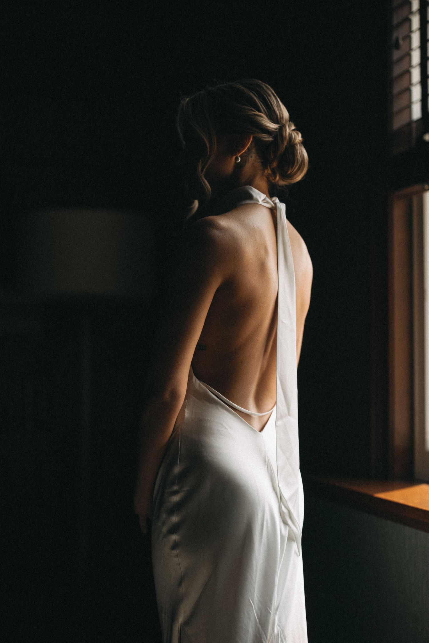 Bride in elegant backless white satin halter gown stands by window in dramatic moody lighting.