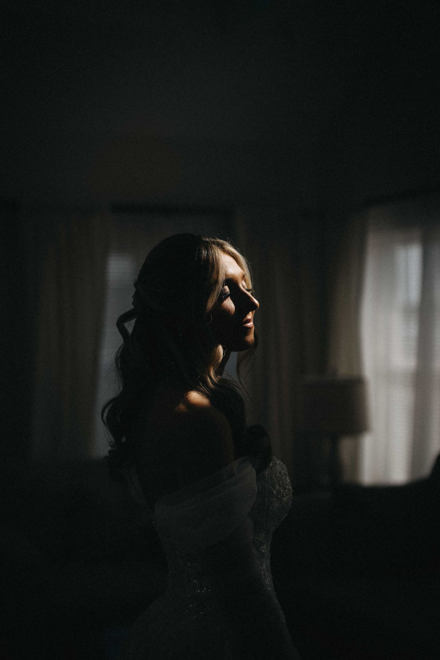 Dramatic moody portrait of a bride in an off-shoulder gown with light and shadow across her face indoors.