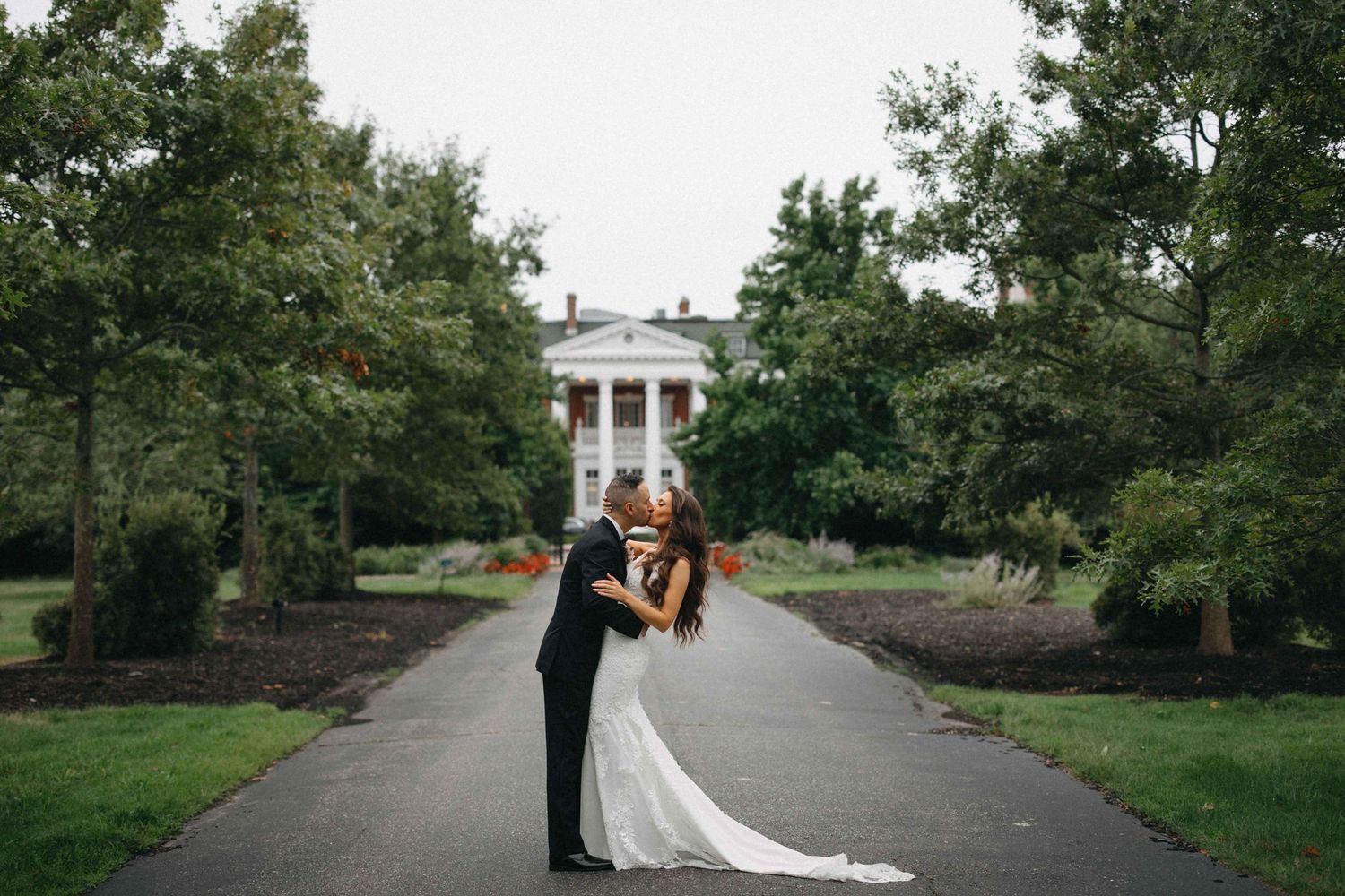 Bride and groom sharing a romantic kiss on a tree-lined path before a grand white Southern mansion.