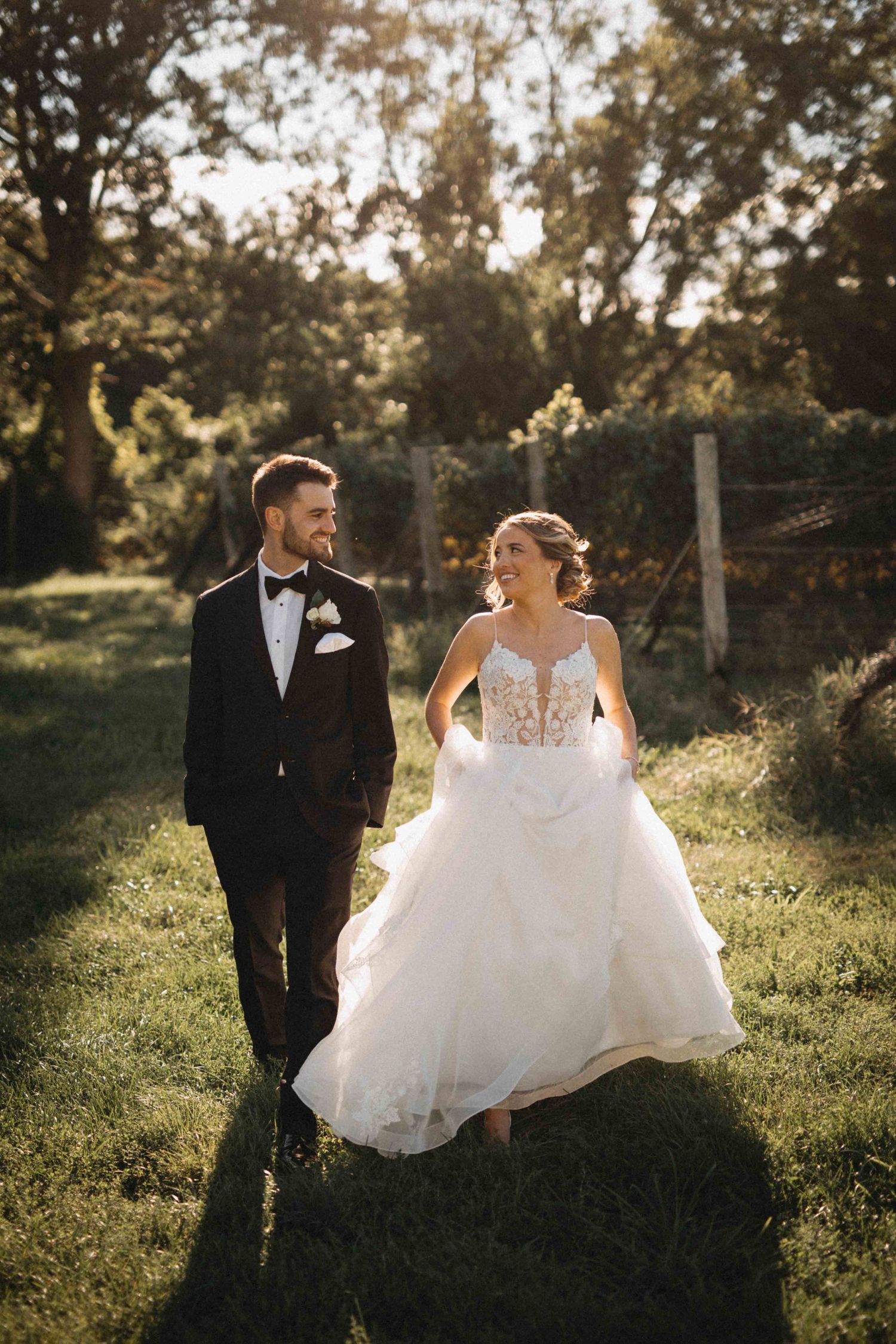 Smiling bride and groom walk hand in hand through sunlit green garden on their wedding day.