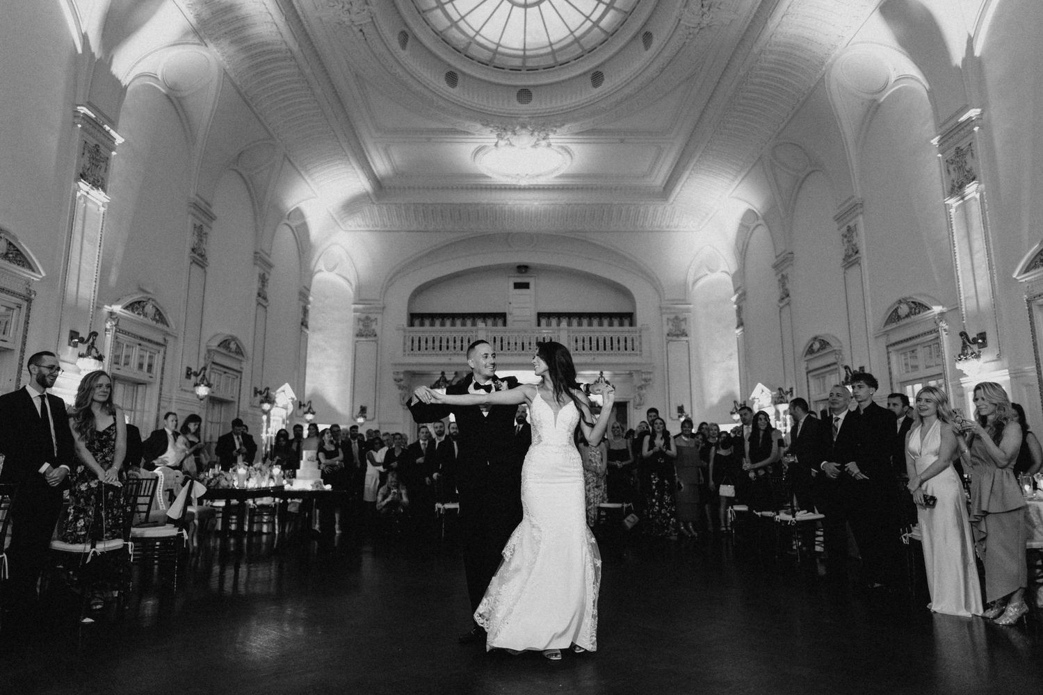 Black and white photo of bride and groom sharing first dance in grand historic ballroom with guests watching.