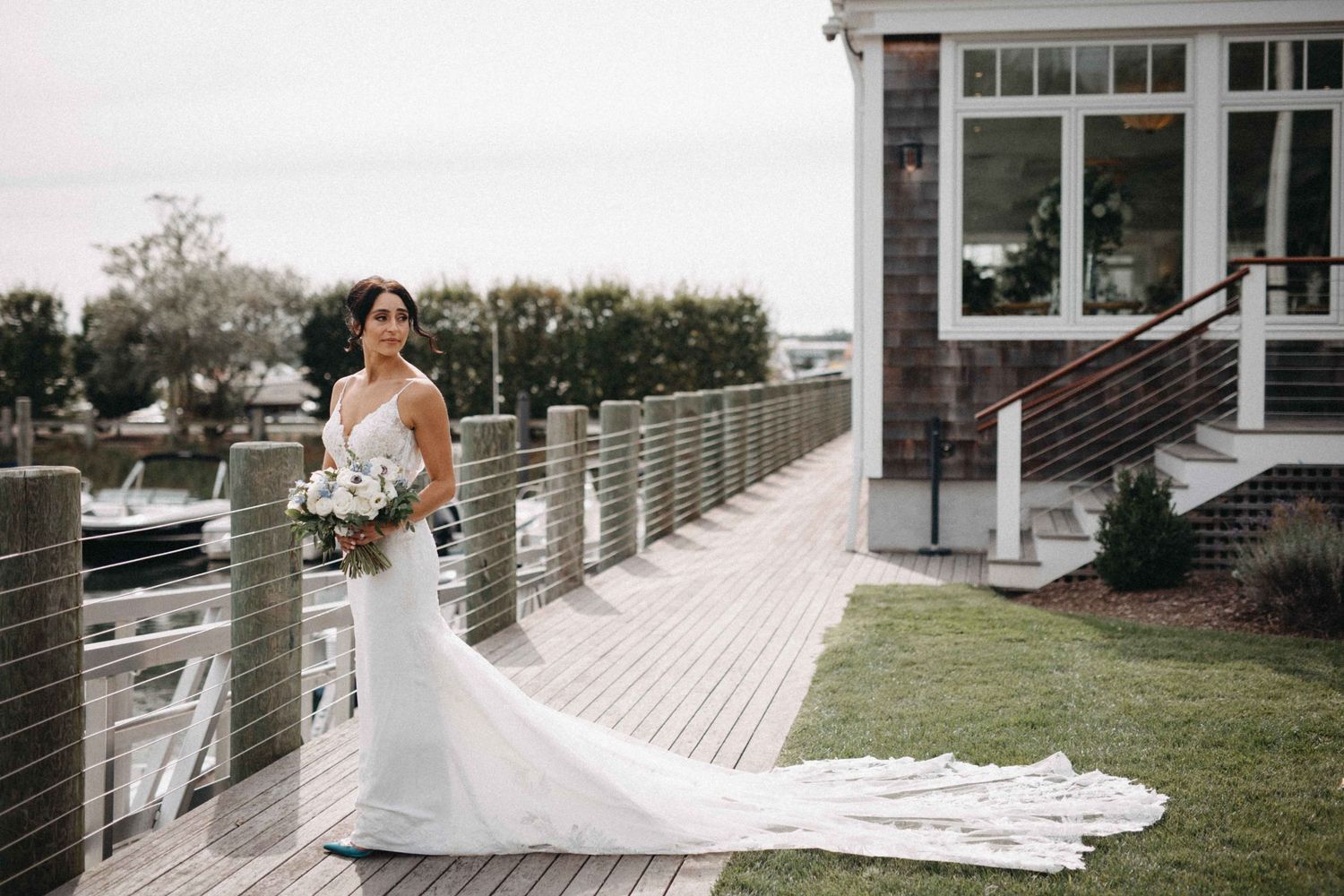 Elegant bride in fitted white gown with long lace train standing on waterfront dock near marina.