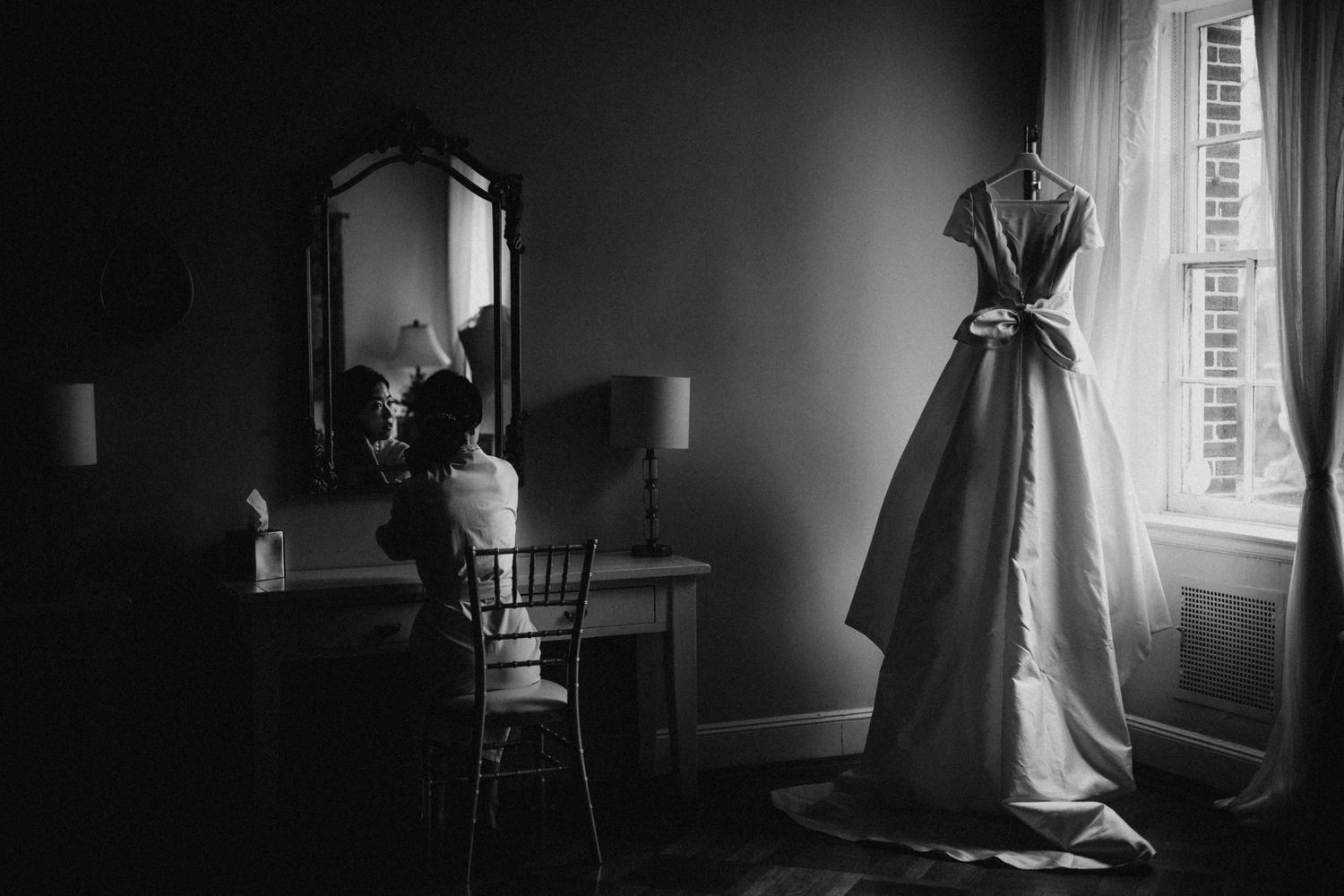 Black and white photo of a bride at a vanity mirror with her gown hanging by the window.
