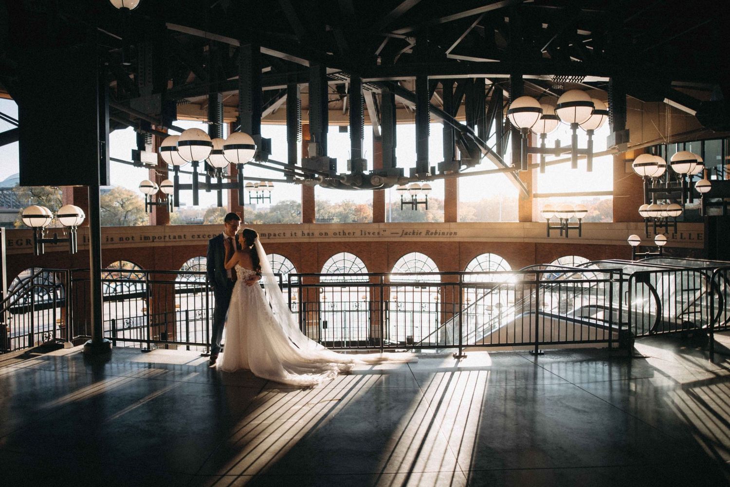 Bride and groom silhouetted against golden sunset light in an industrial venue with arched windows.