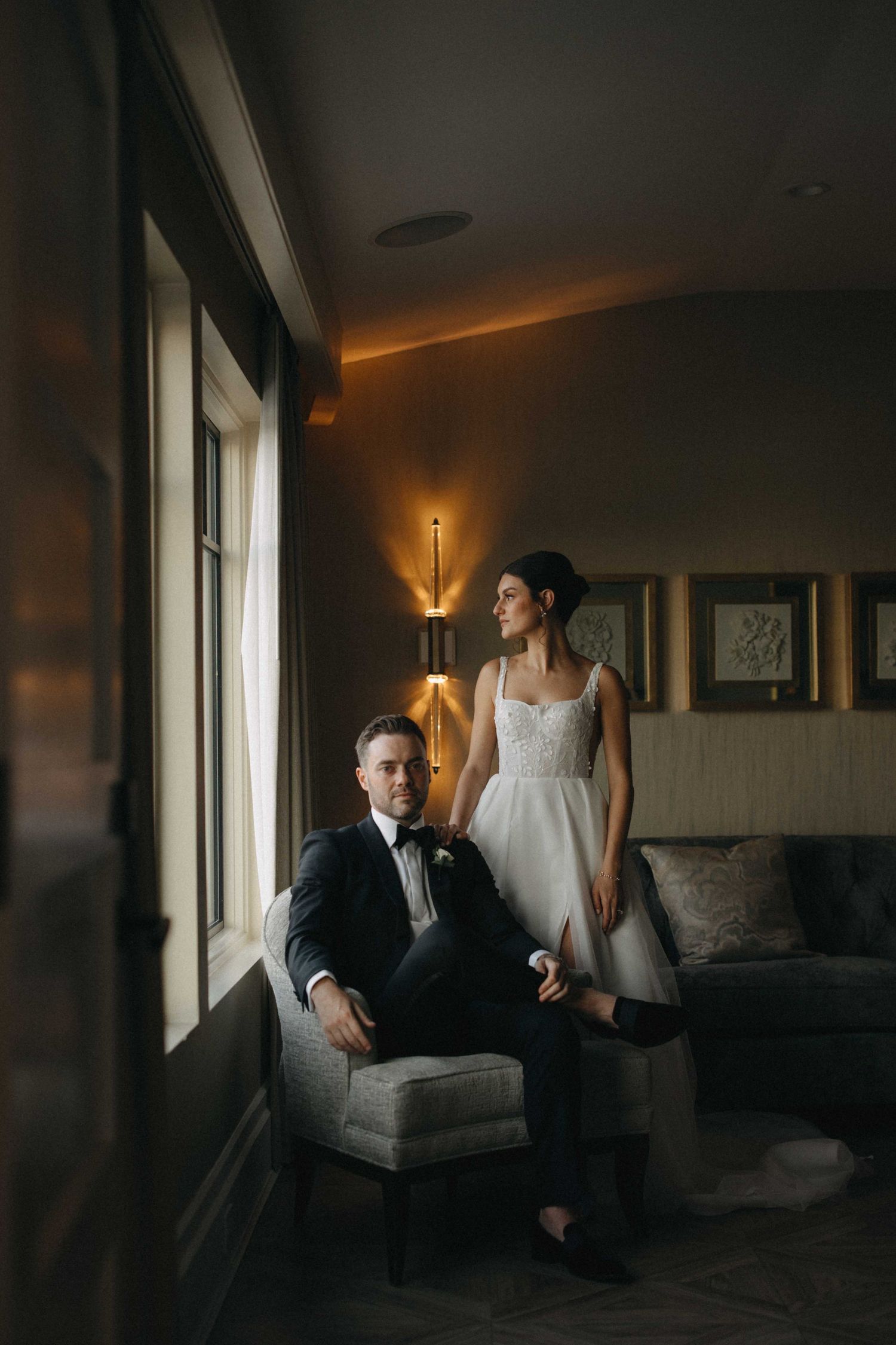 Bride and groom posing in a warmly lit hotel suite, groom seated by window as bride stands beside him.