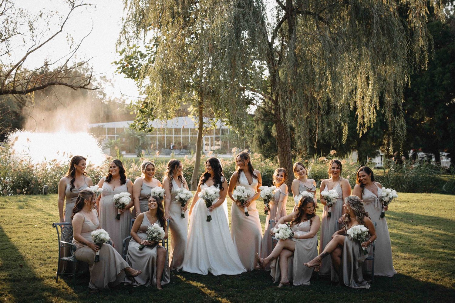 Bride surrounded by bridesmaids in taupe dresses holding white bouquets under weeping willow by lake.