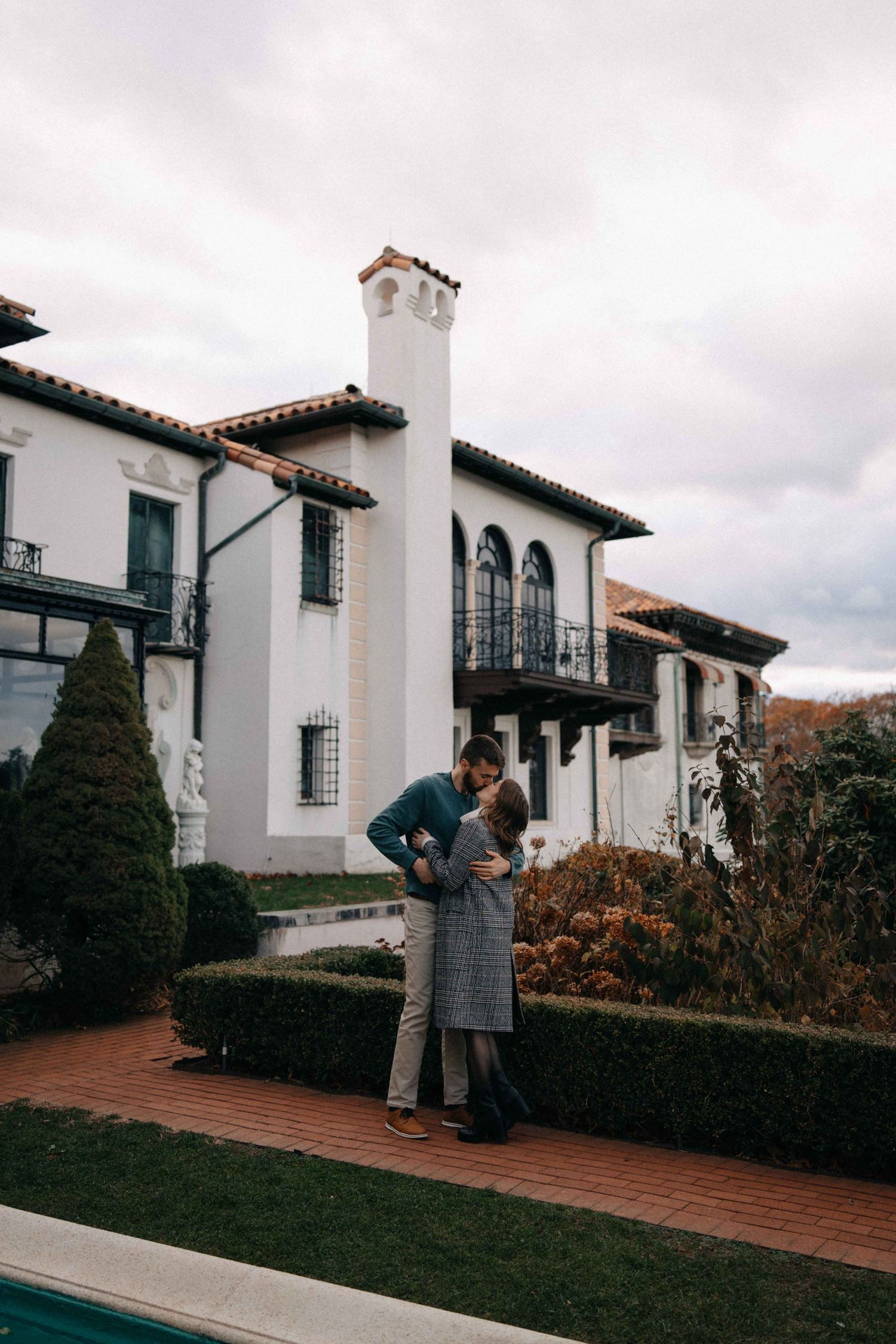 Couple embracing in front of a white Spanish-style villa with manicured hedges.