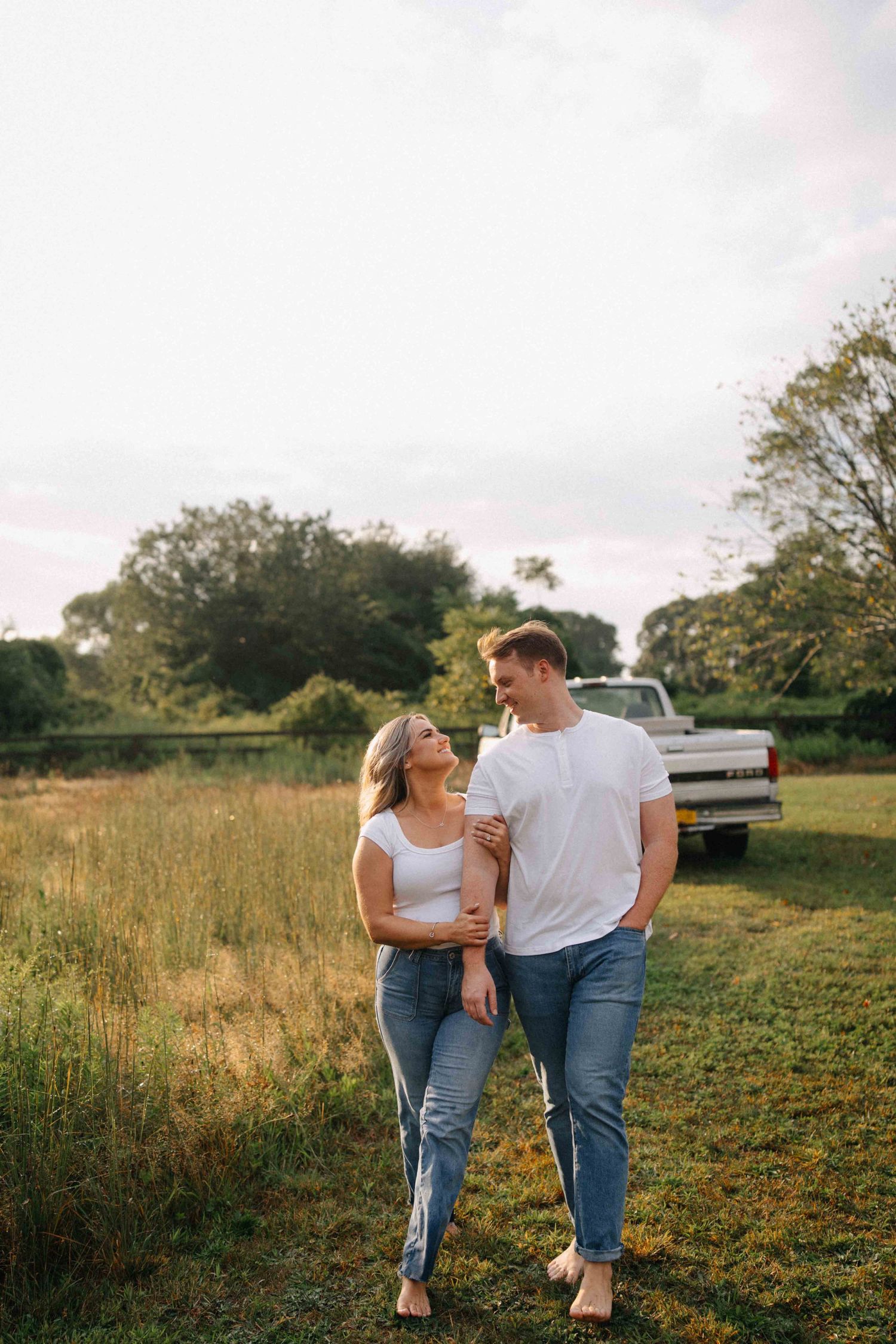 Couple walking barefoot together through a golden meadow with a vintage white pickup truck behind them.