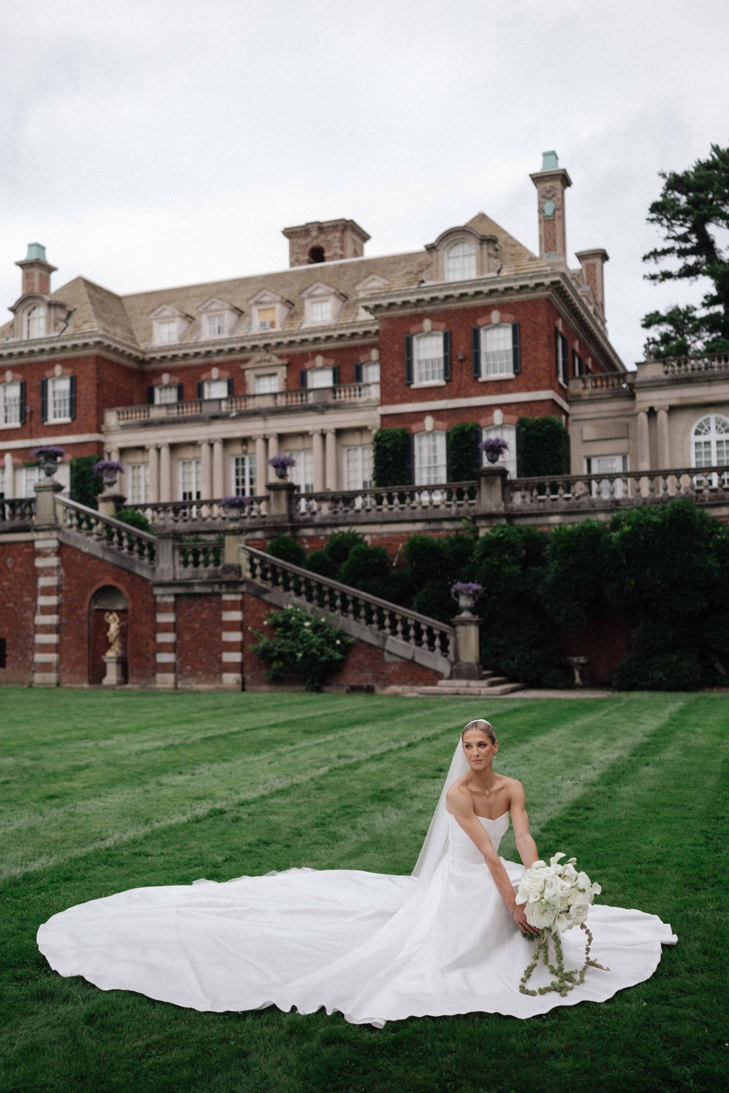 Bride in a white gown with long veil sits on green lawn before a grand red brick mansion.