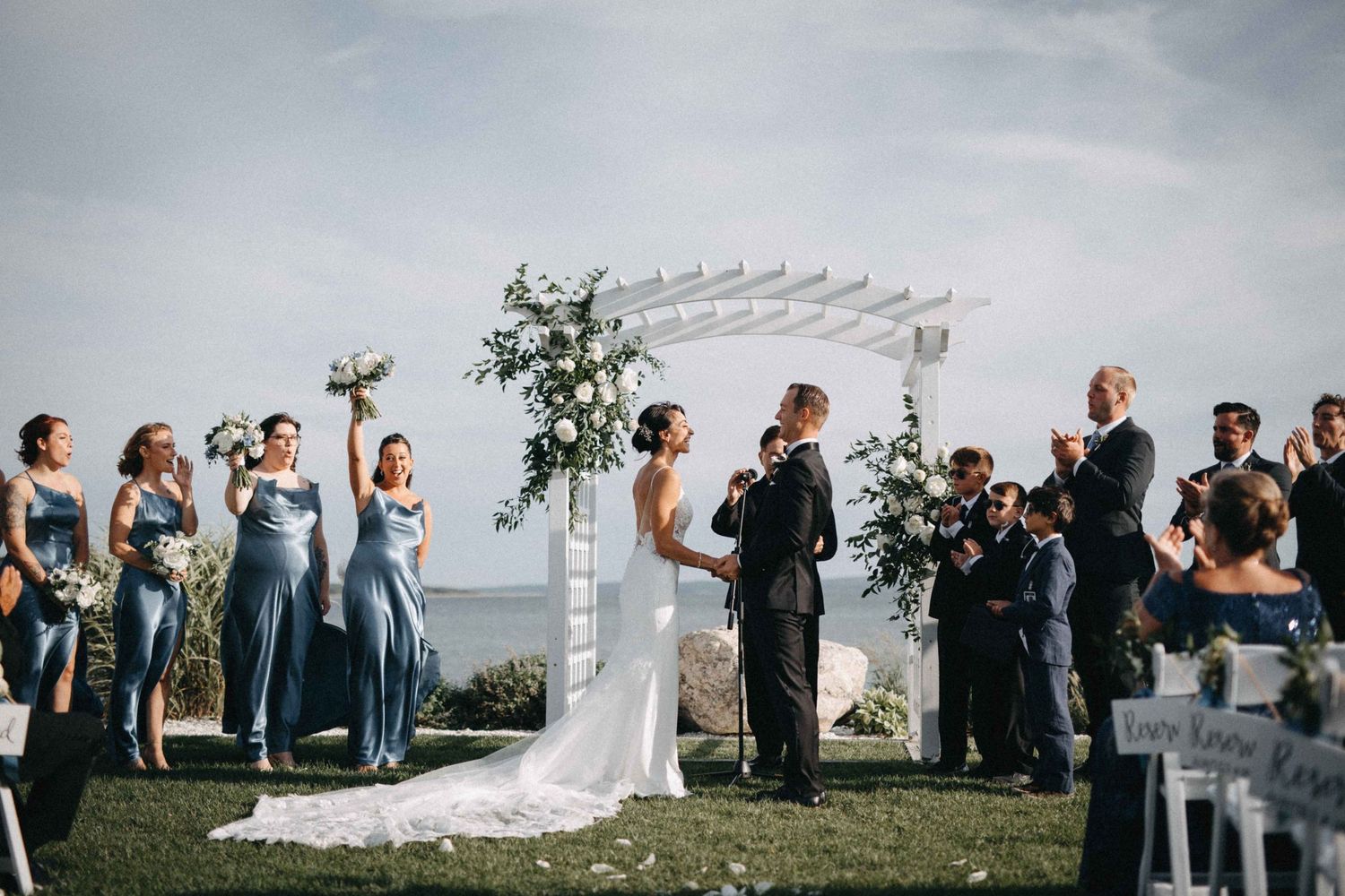 Outdoor waterfront wedding ceremony with bride and groom at white arbor surrounded by guests and bridesmaids.