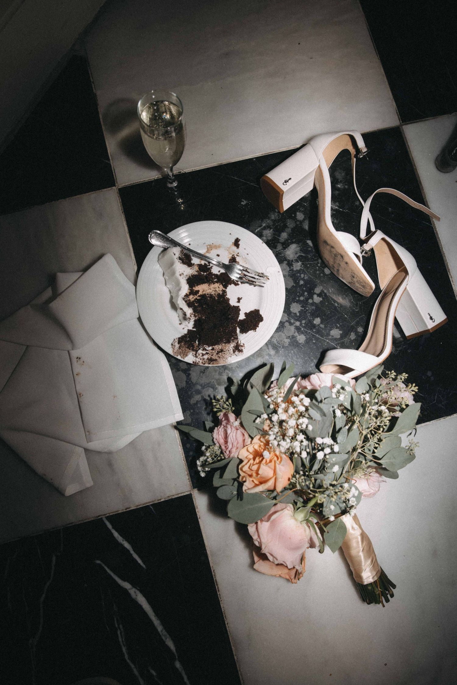 Flat lay of bridal heels, bouquet, champagne flute, and half-eaten cake slice on checkered marble floor.
