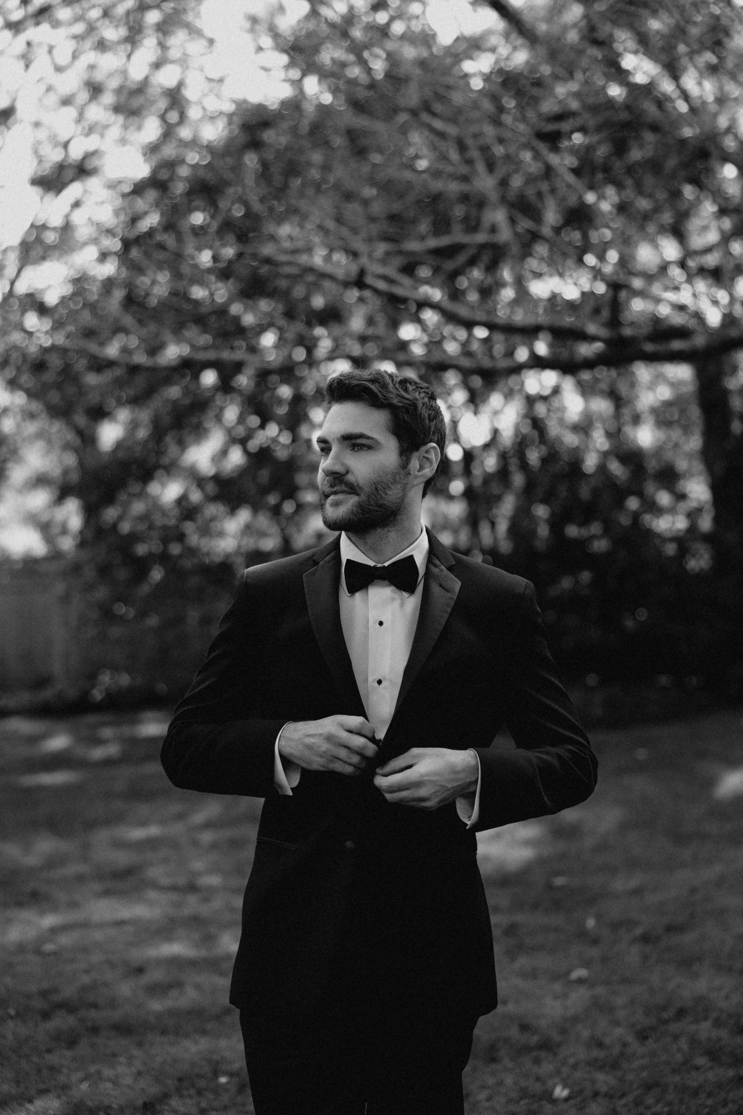 Handsome groom in black tuxedo and bow tie buttoning jacket in black and white outdoor portrait.