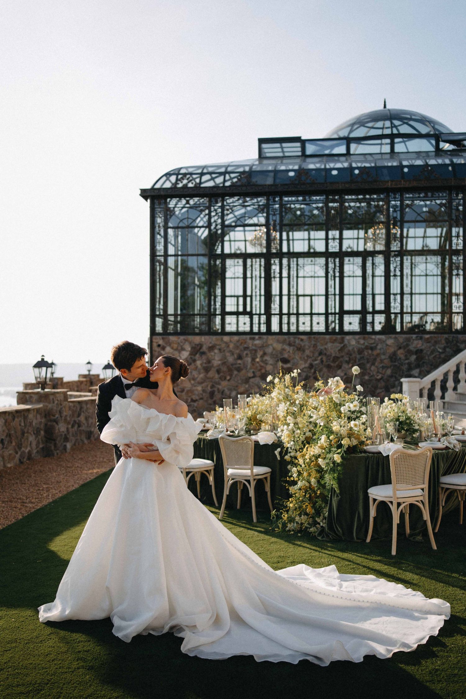 Bride and groom share a romantic kiss at an elegant outdoor wedding venue with a glass greenhouse backdrop.