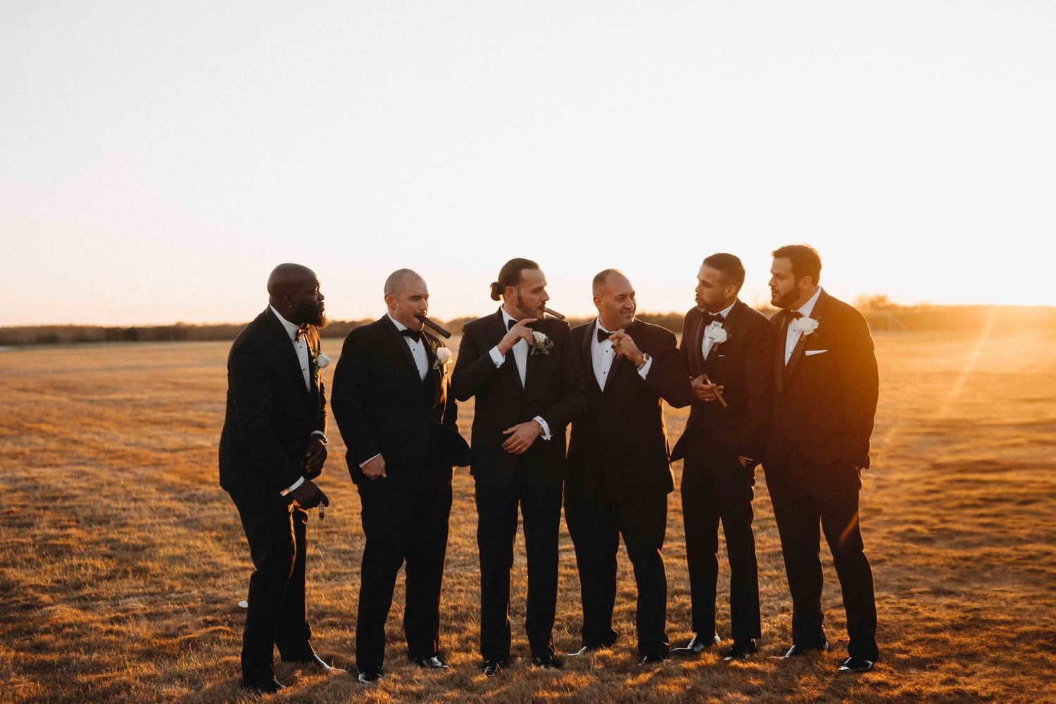 Groom and groomsmen in black tuxedos laughing together in a golden sunset field.