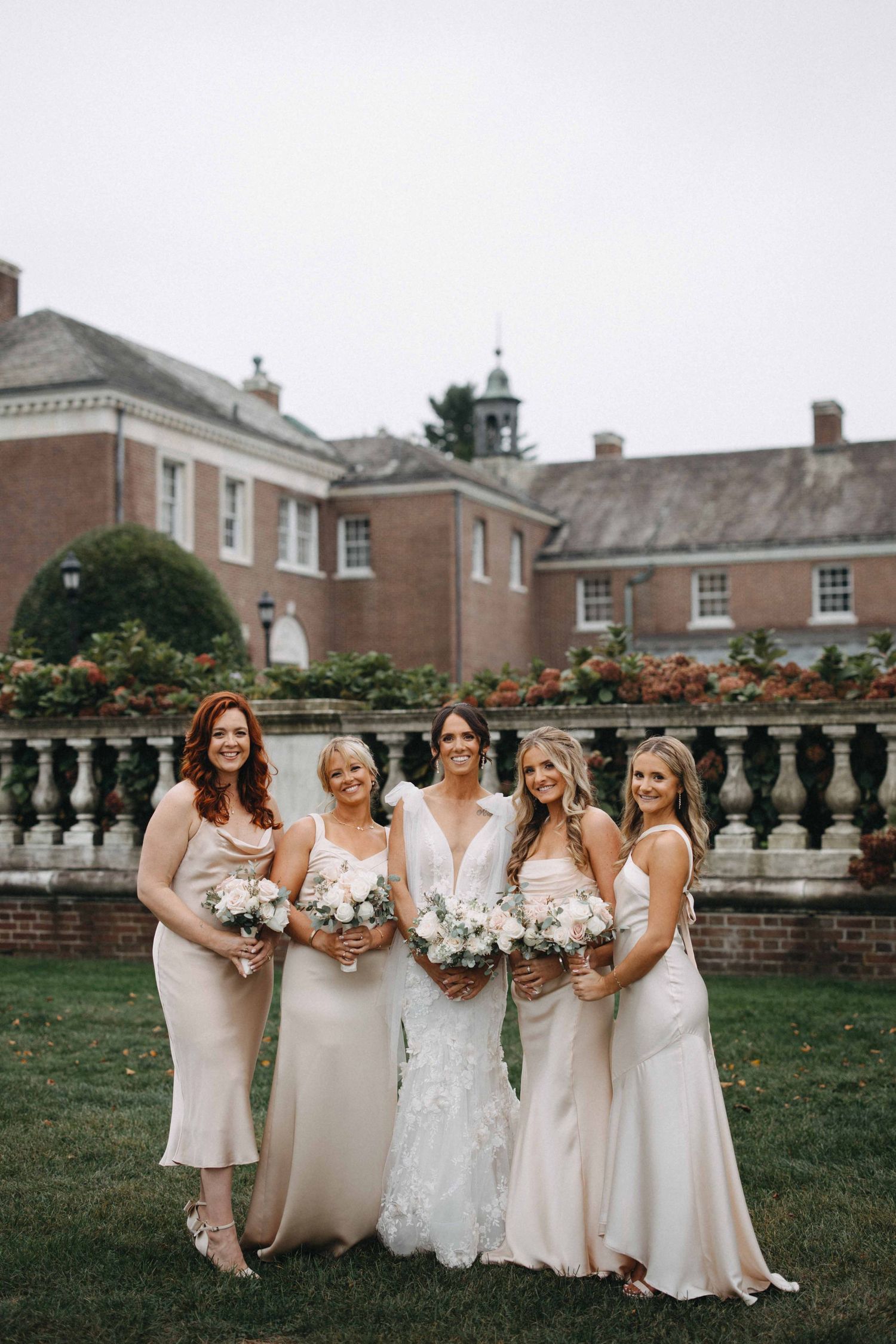 Bride with four bridesmaids in champagne satin dresses holding white bouquets on lawn before brick estate manor.