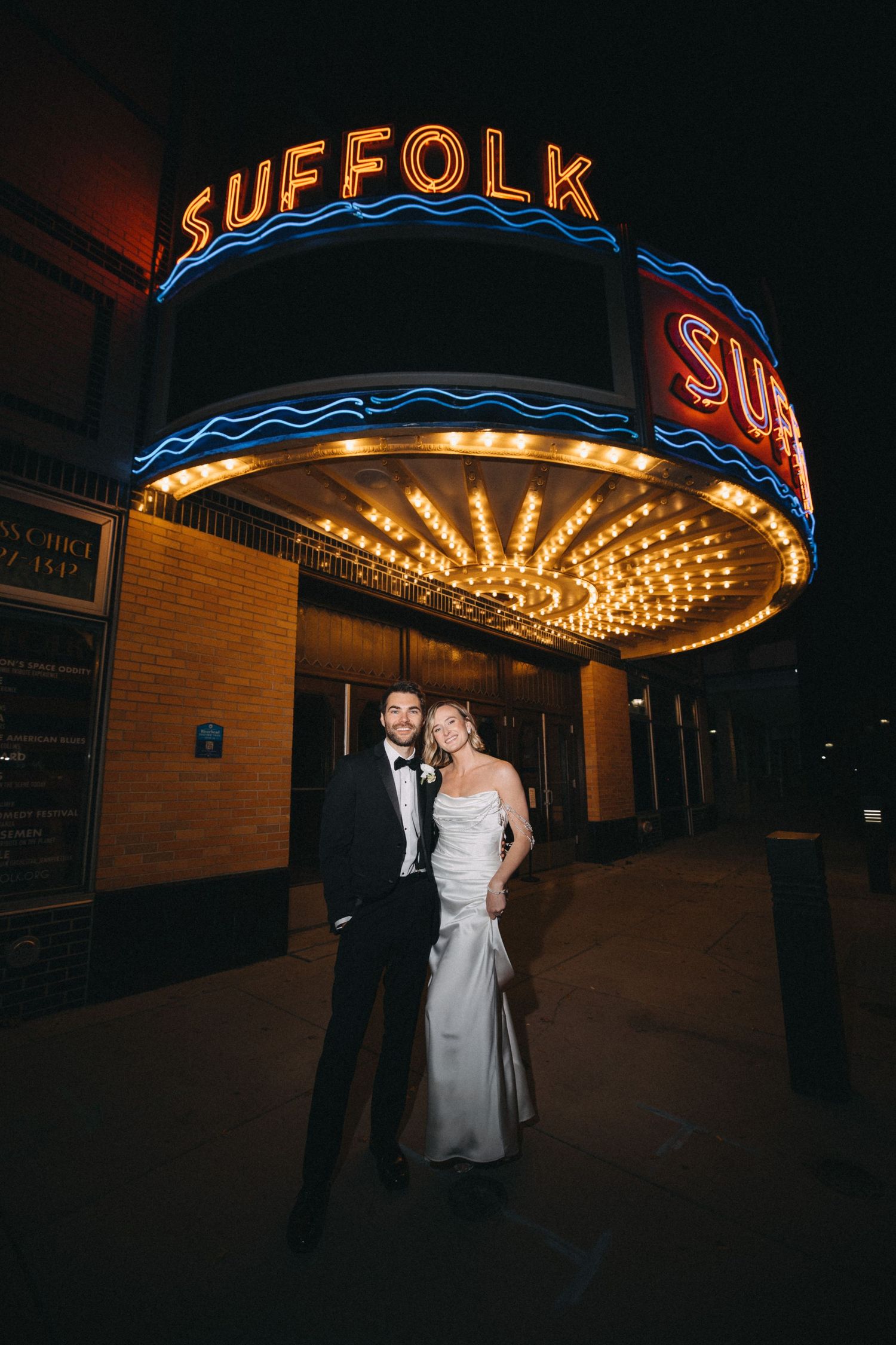 Bride and groom pose under the glowing Suffolk Theater marquee sign at night.