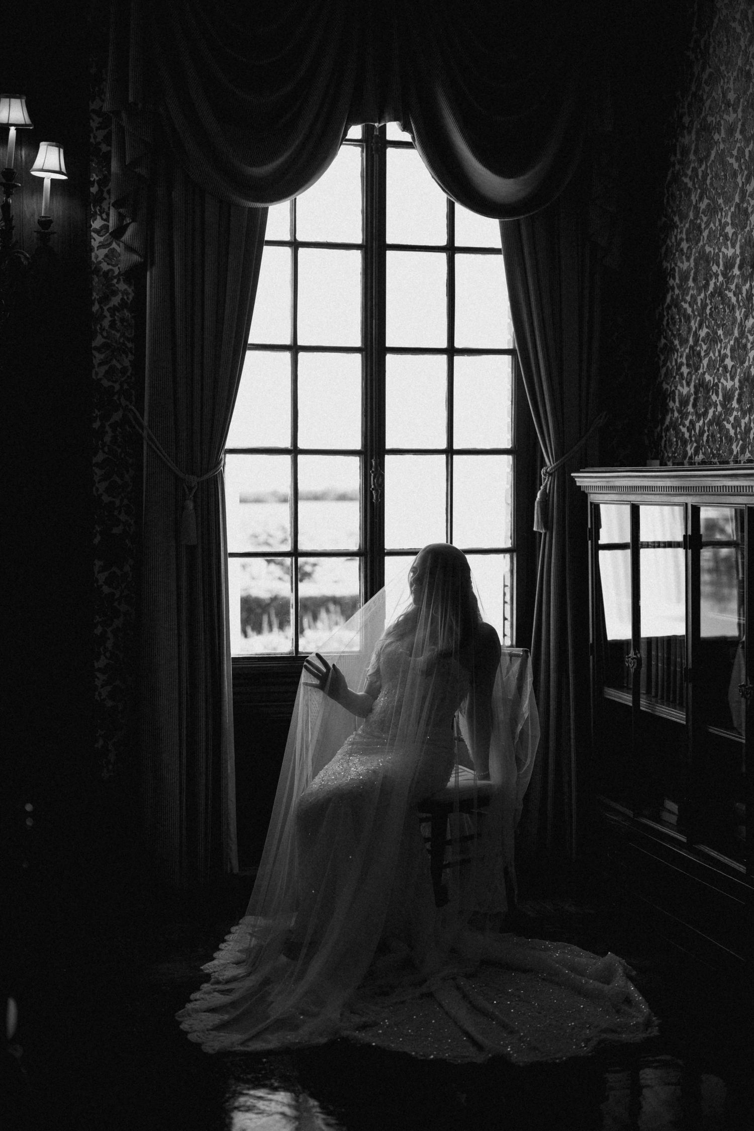 Black and white photo of a bride in wedding dress and veil sitting by large window in elegant dark room.