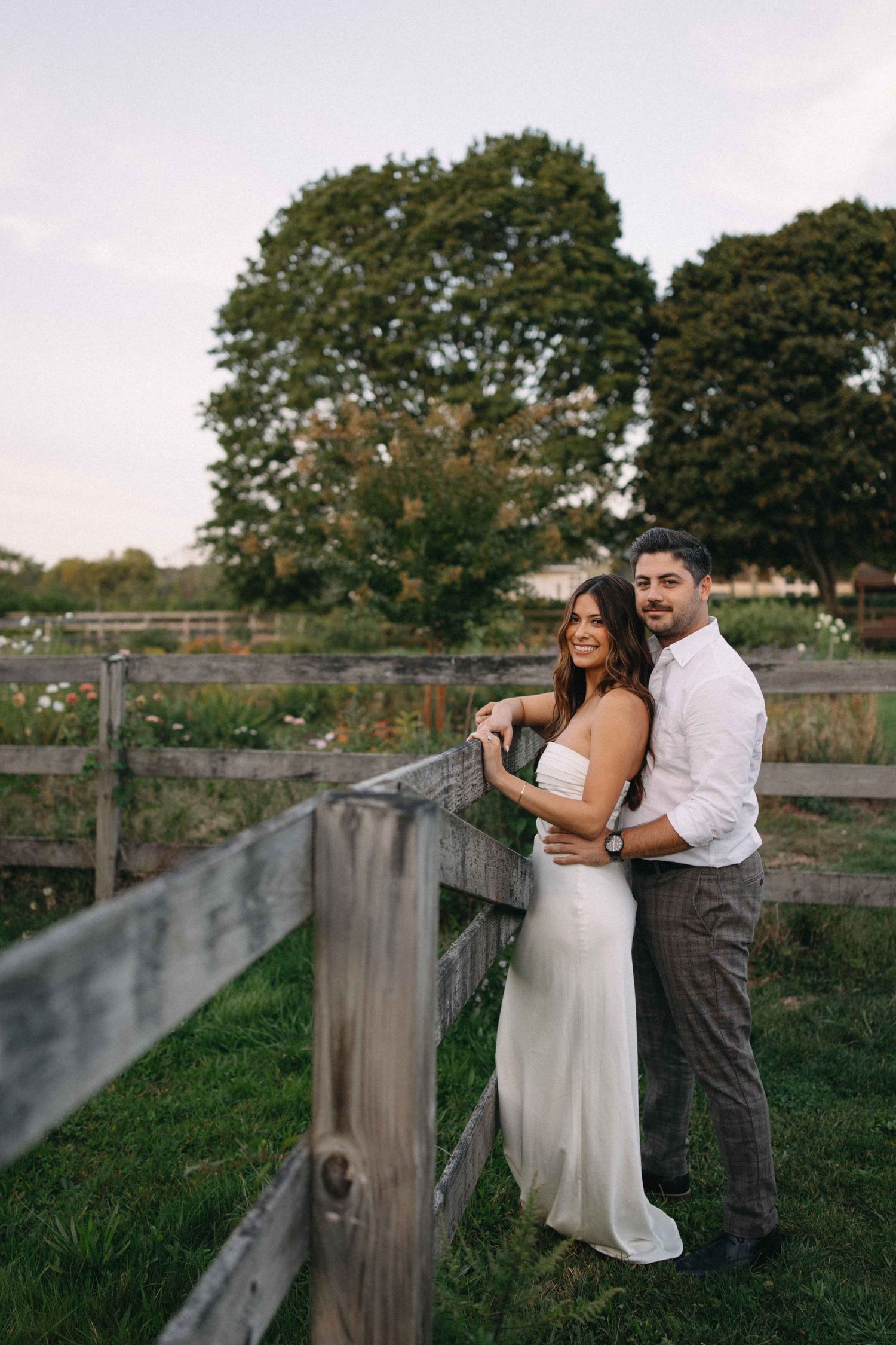 Couple embracing by rustic wooden fence at farm, woman in white dress, man in white shirt at dusk.