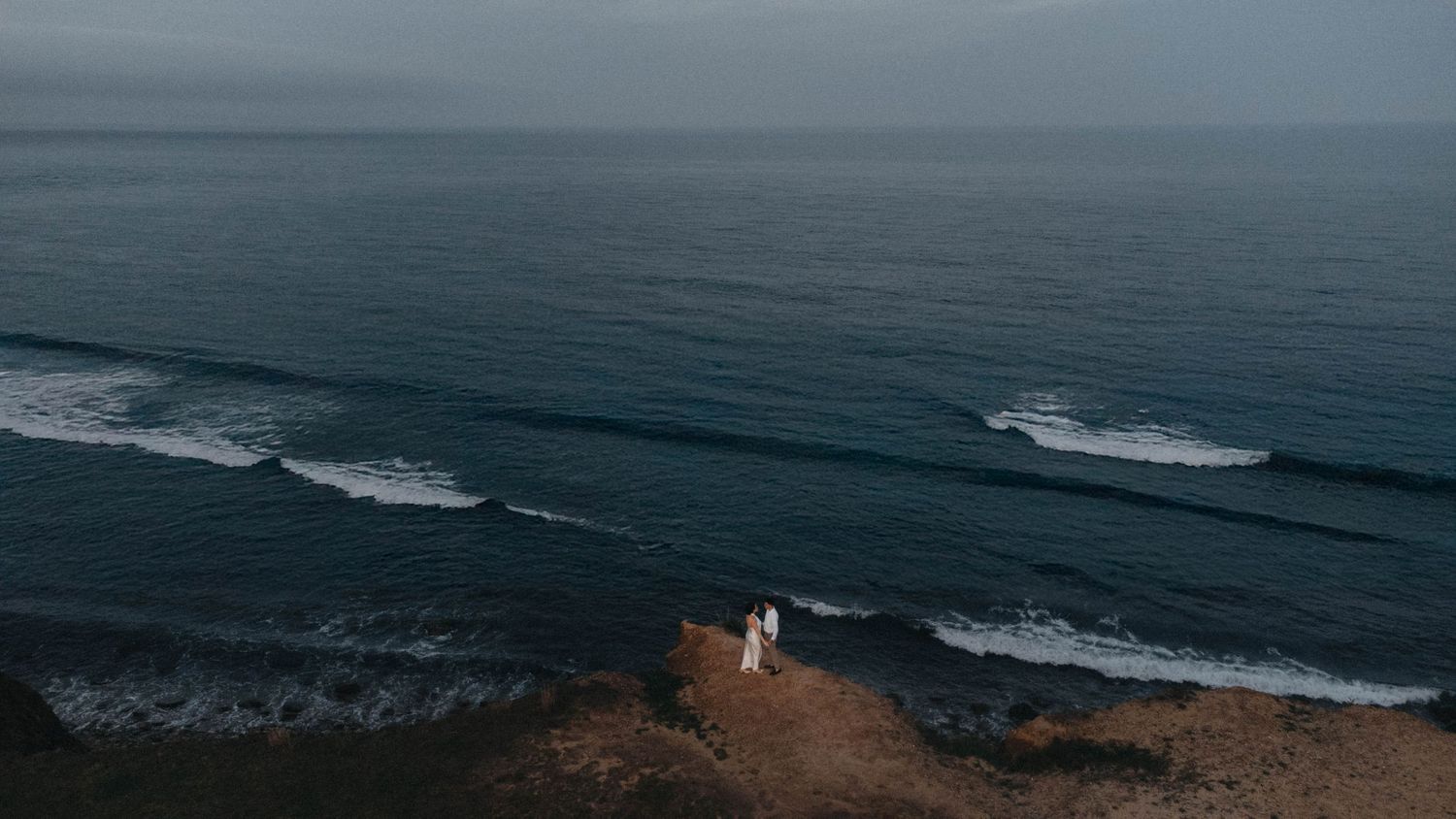 Aerial drone shot of bride and groom standing on coastal cliffs above ocean.