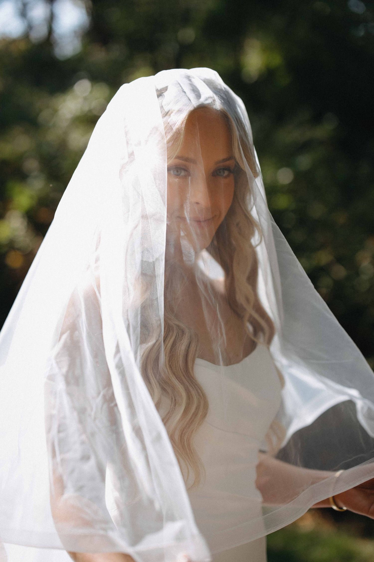 Blonde bride with wavy hair draped in sheer white veil looking at camera outdoors.