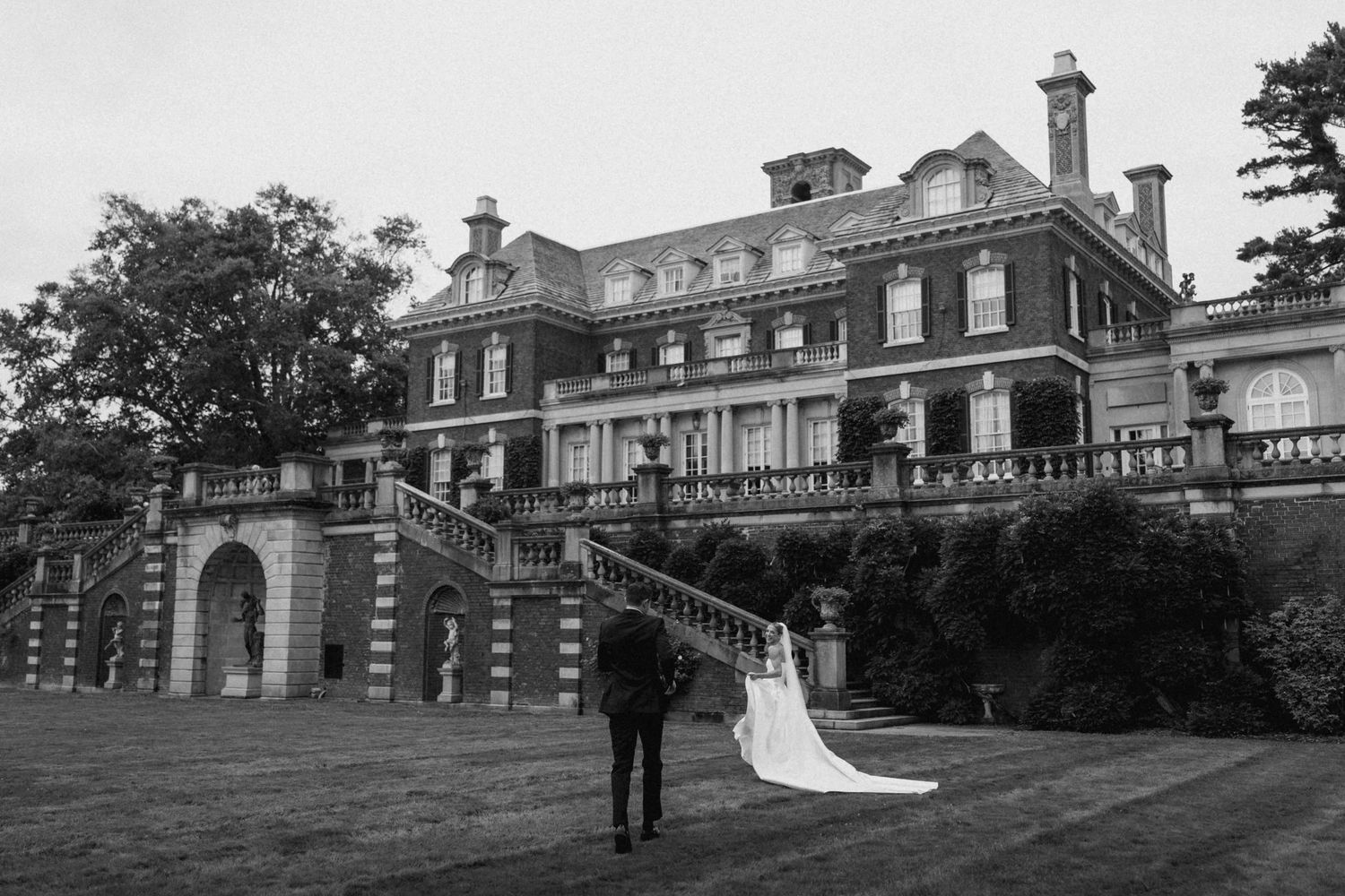 Black and white wedding photo of couple standing before a grand historic brick mansion with elegant staircases.