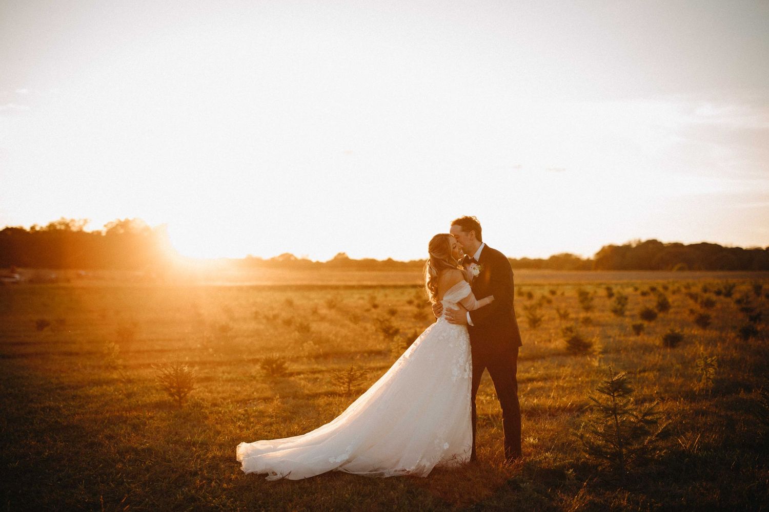 Bride and groom embracing in a sun-drenched field with stunning golden hour sunset backdrop.