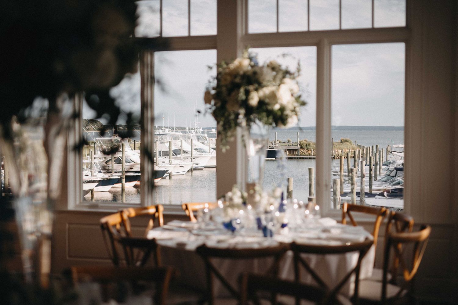 Wedding reception tables with tall white floral centerpieces framed by large windows overlooking a marina.